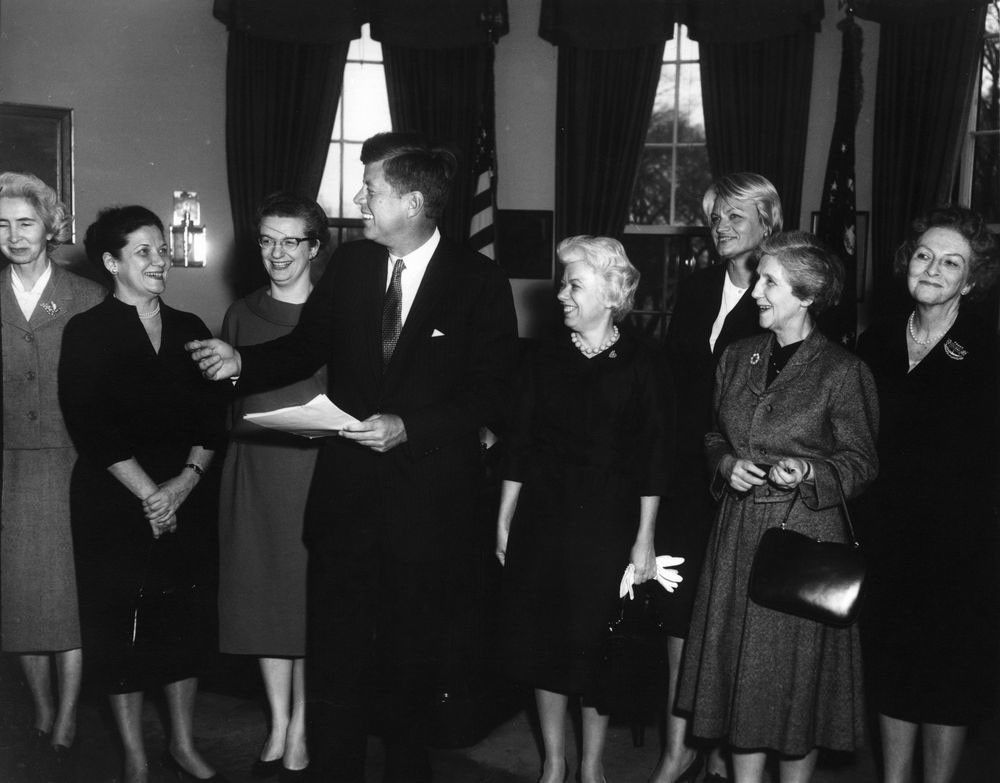 Nancy Grace Roman with President John F. Kennedy and six other women in the Oval Office. Roman is the third from the left, standing next to Kennedy. Kennedy is speaking to the woman to the left of Roman.