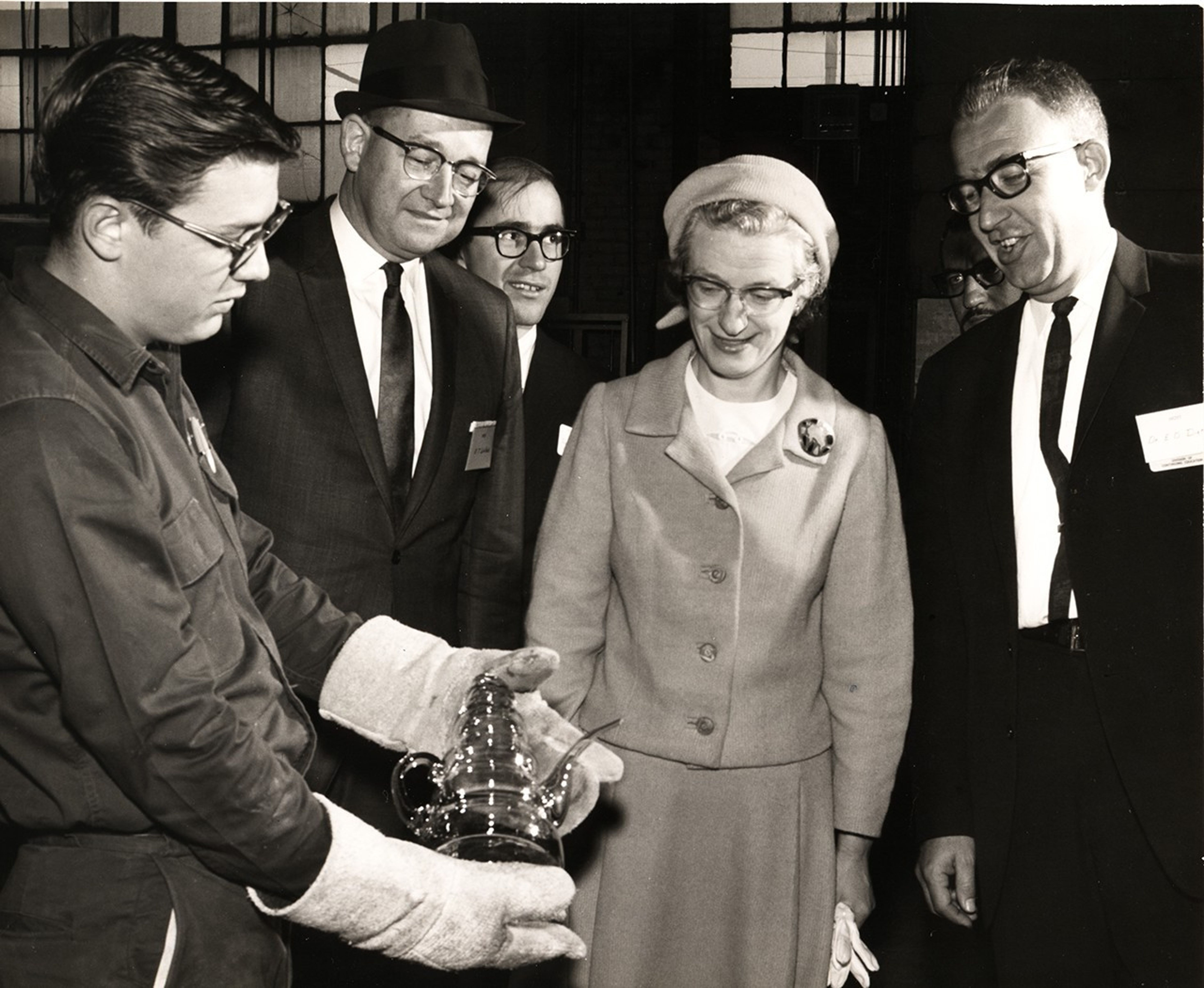 Nancy Grace Roman stands in the center of a group of five men, one of whom wears thick gloves and presents her with a glass teapot.