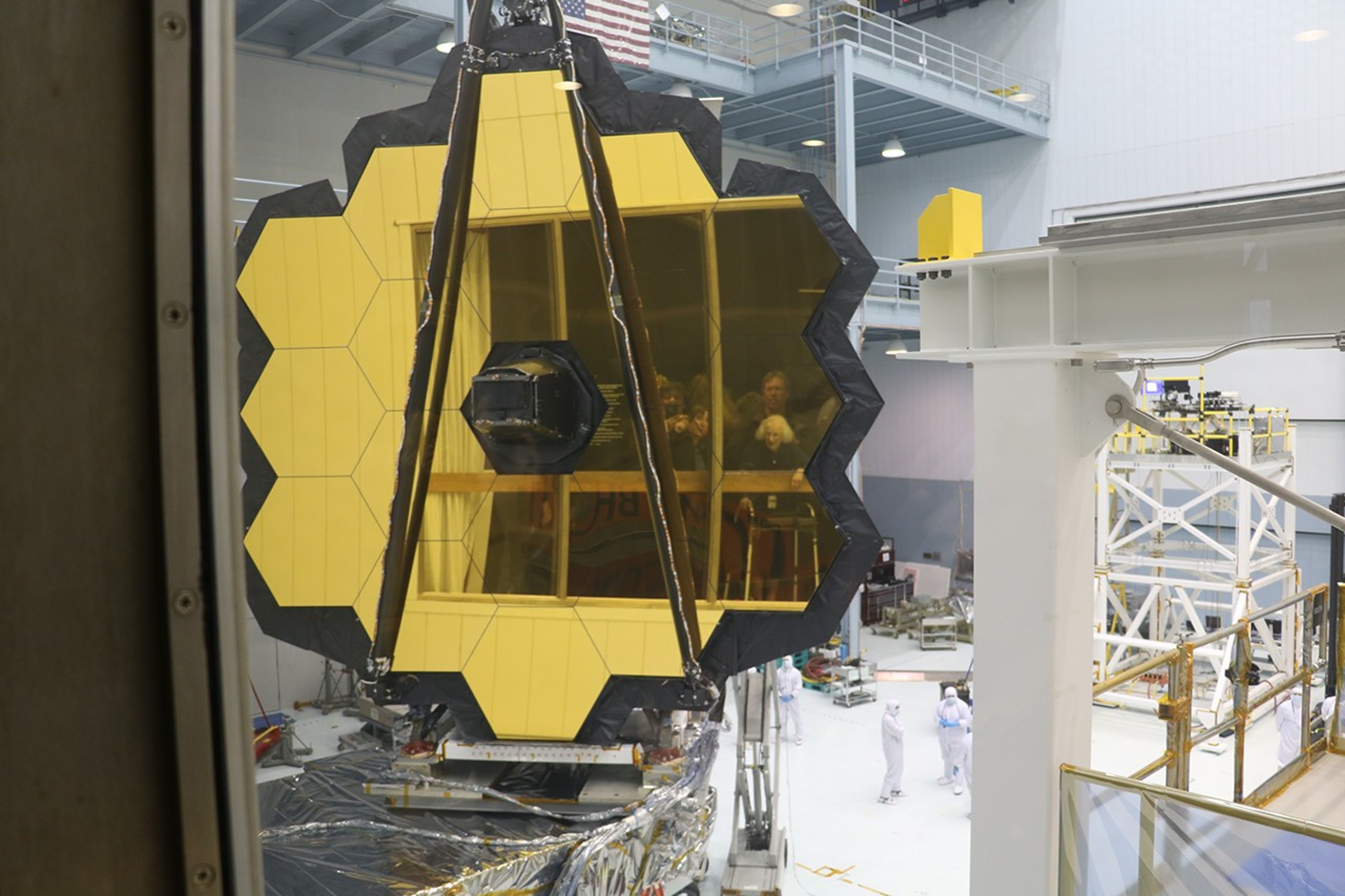 The James Webb Space Telescope is being assembled in the GSFC clean room. In its mirrors, the reflections of Dr. Roman, Dr. Rigby, and other visitors are visible.