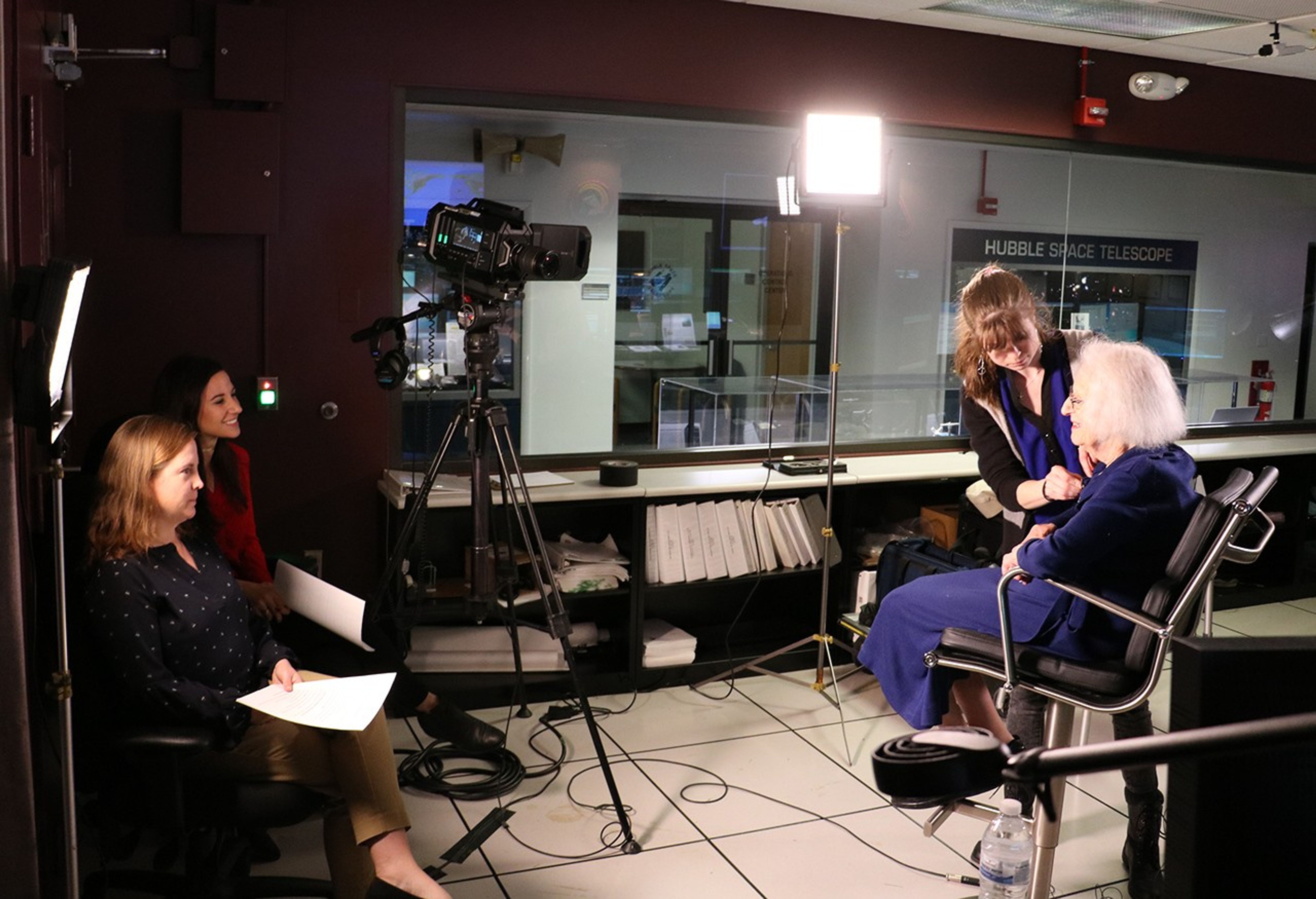 Dr. Roman sits in front of the camera, prepared for the interview, as Katrina Jackson attaches a microphone to her shirt. Morgan Van Arsdall and Erin Kisliuk wait nearby to begin the interview.