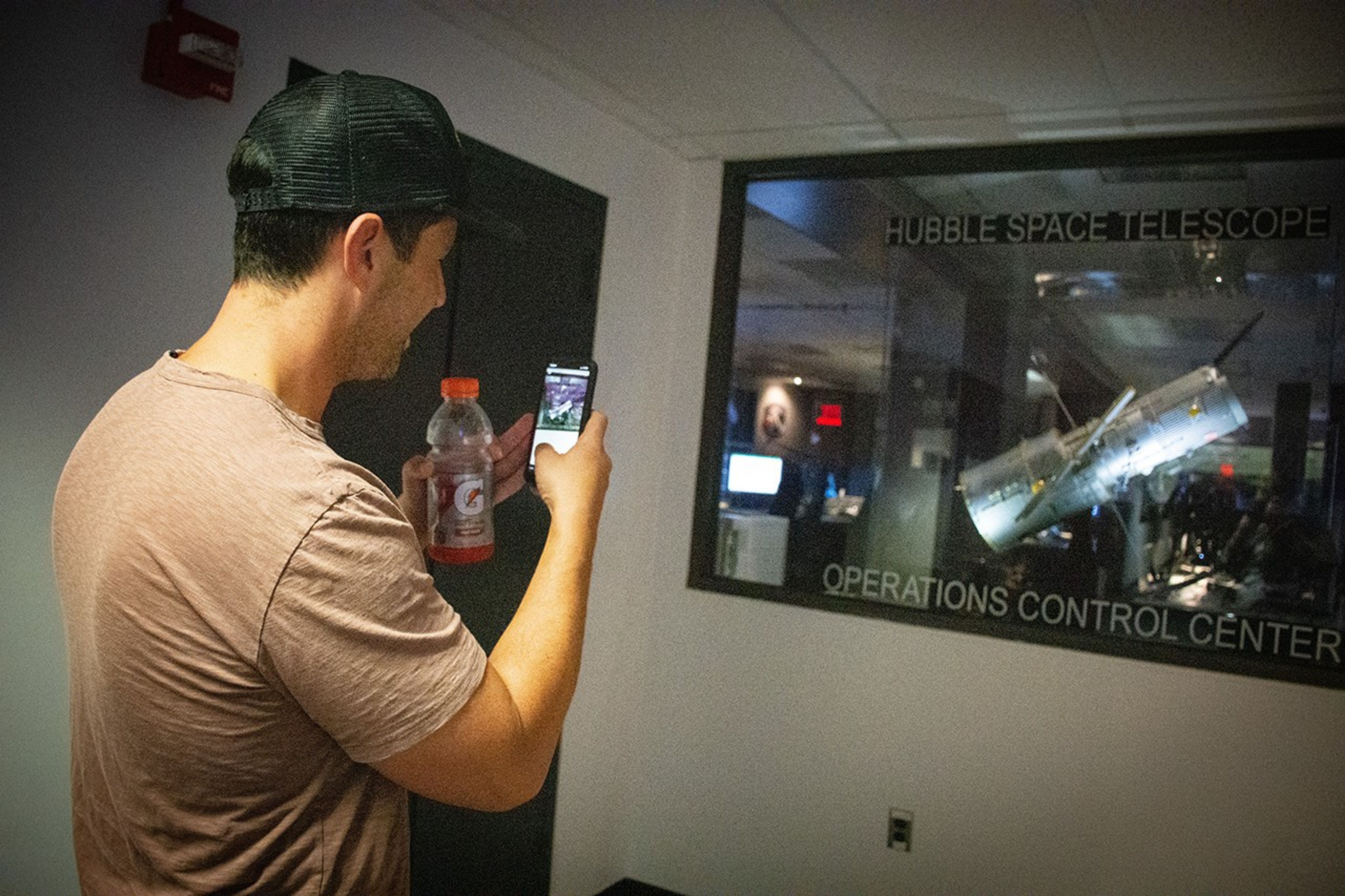 Peck, in the foreground, faces away from the camera while pointing his smartphone at the clear glass window of the Operations Control Center to take a picture.