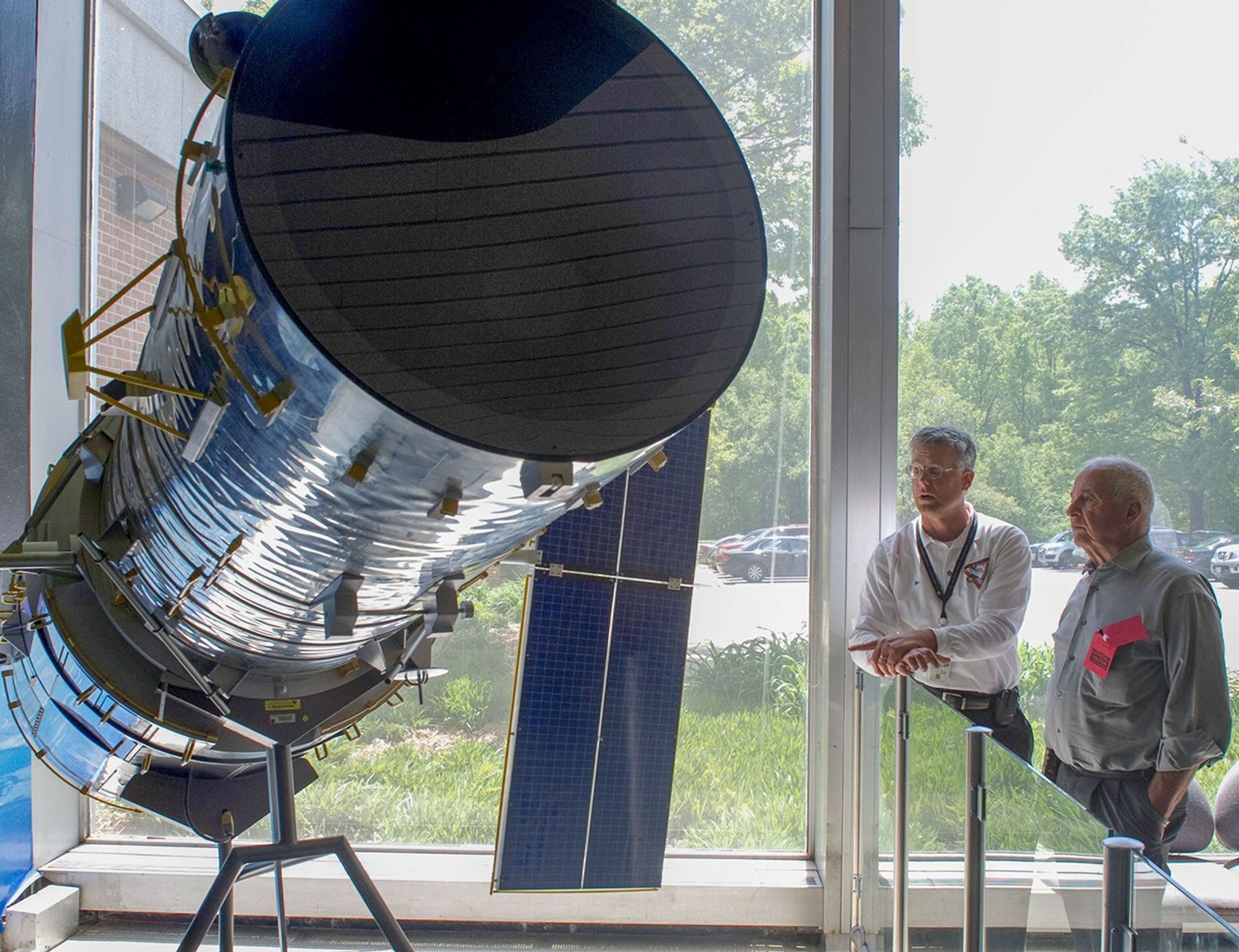 Jim Jeletic and Bill Anders stand to the right side of the photo, with Jeletic (on the left) talking to Anders (on the right). On the left side of the image is the Hubble model. In the background, through clear glass window's, it's a sunny day with a parking lot, green grass, and lots of trees.