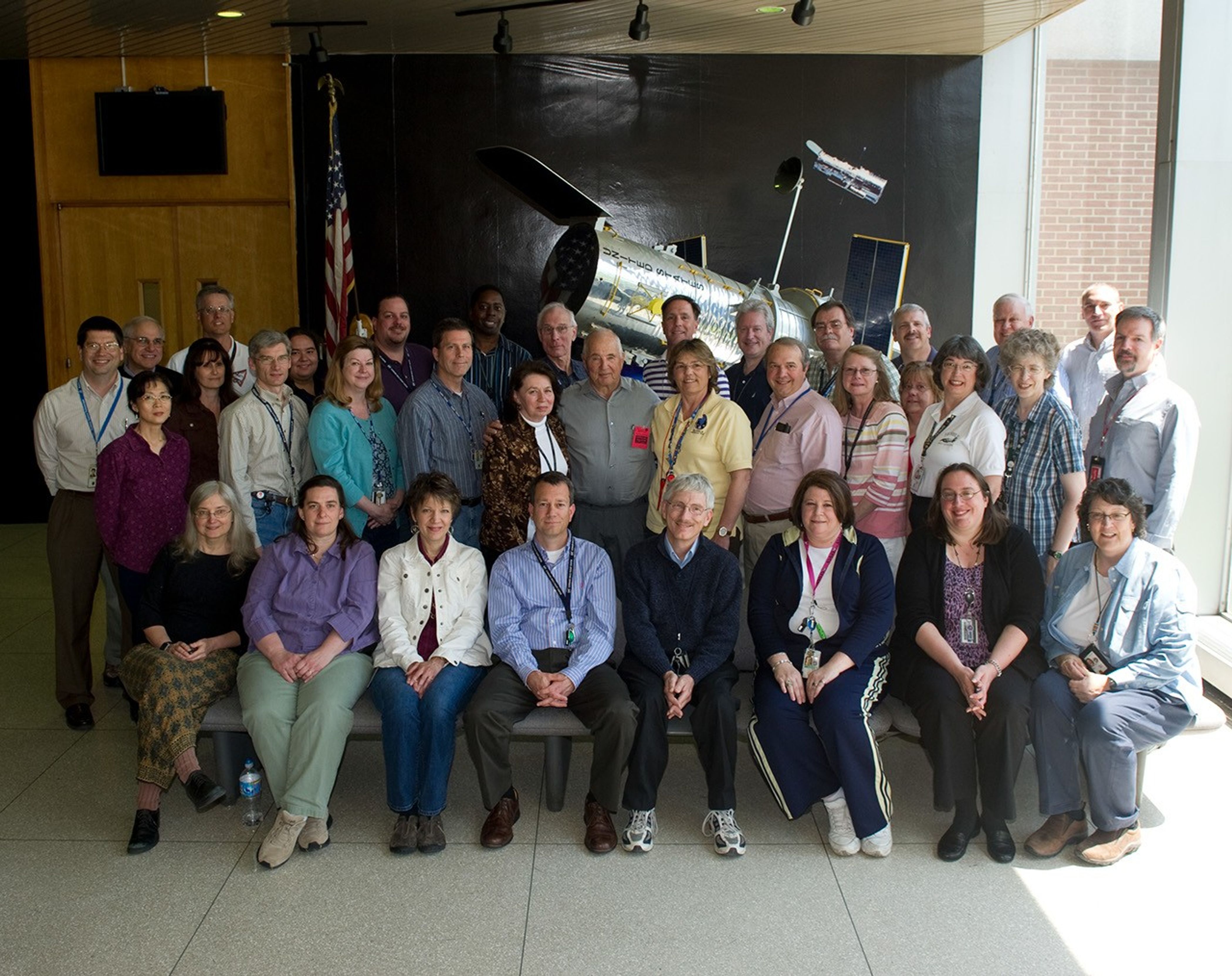 Astronaut Bill Anders stands in the center of a group of 35 people in 3 rows, standing in front of a 1/5 scale model of Hubble.