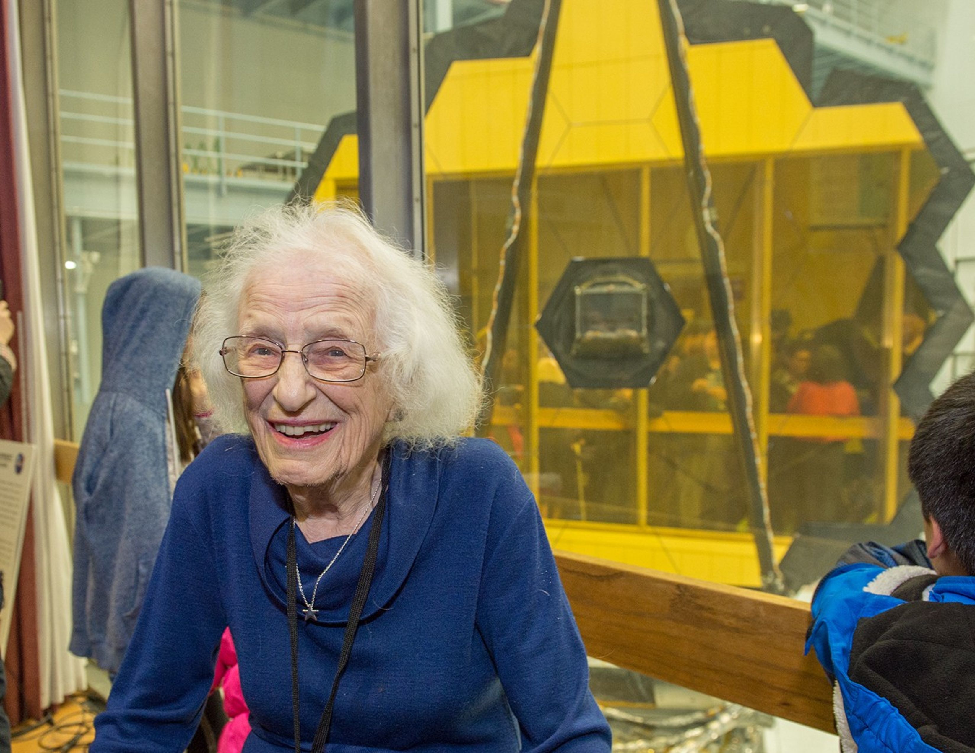 Dr. Roman faces the camera, while in the background, kids view the clean room where JWST is being assembled