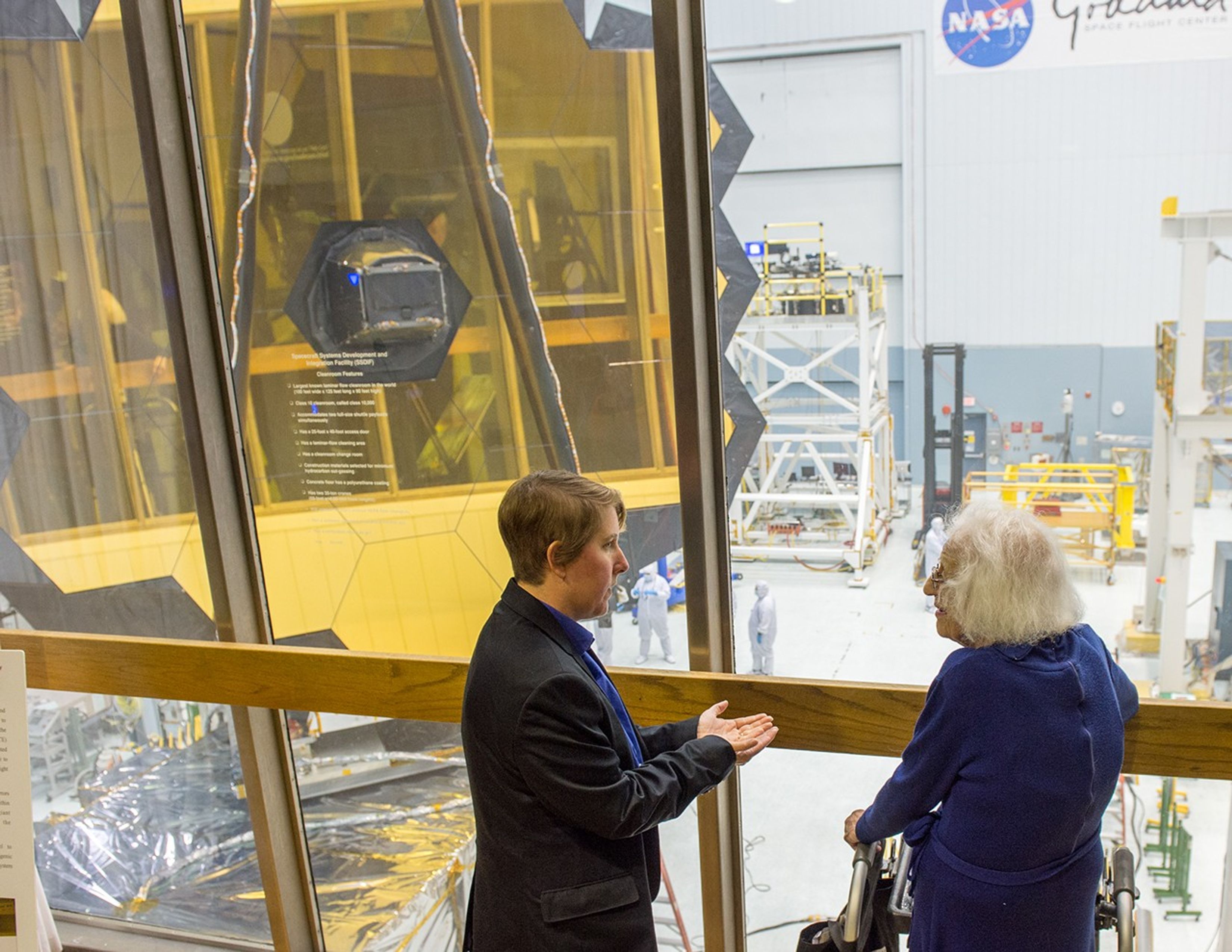 Dr. Rigby, left, cups her hands while speaking to Dr. Roman, right. In the background is the clean room where the James Webb Space Telescope is being assembled, with the telescope on the left side of the image.