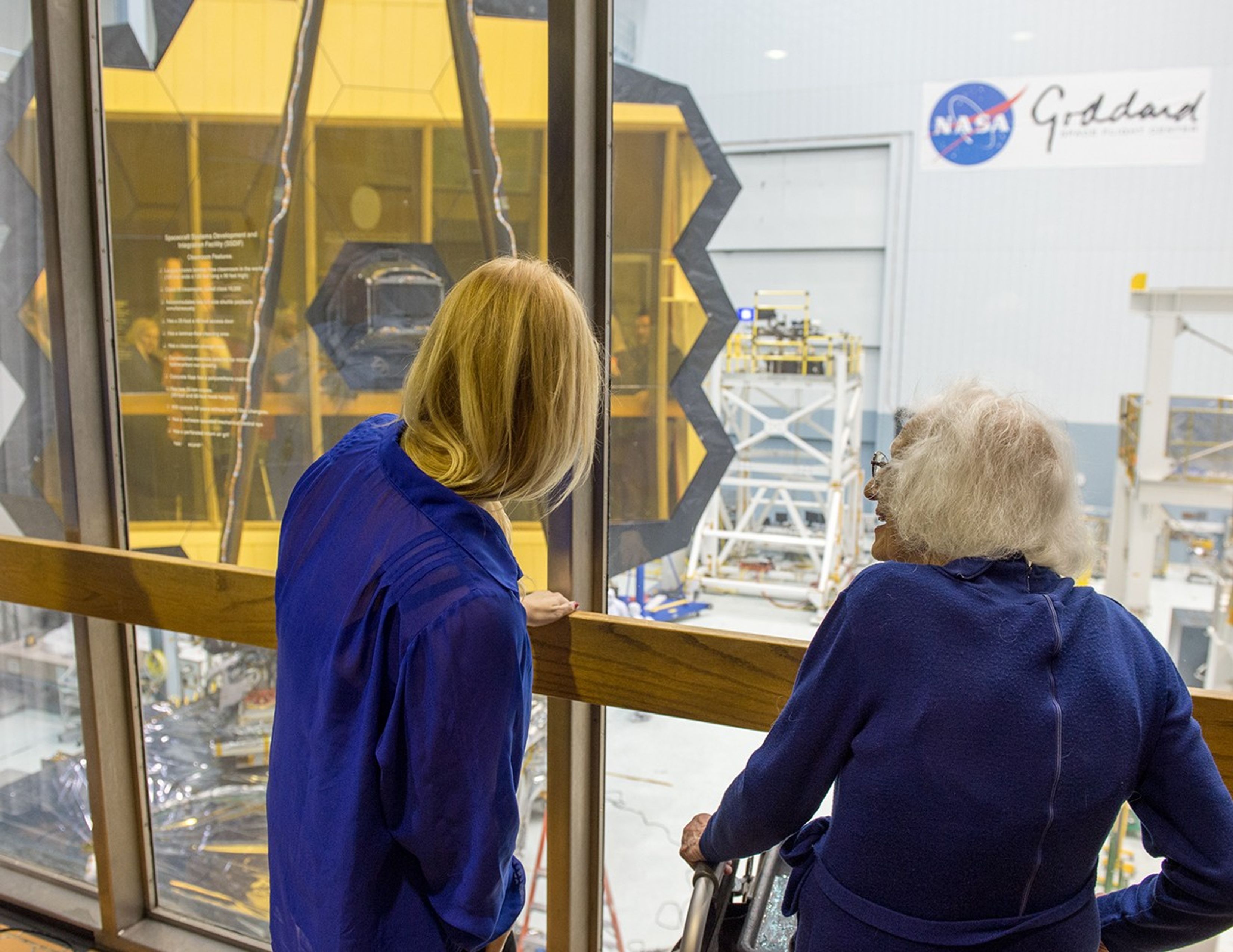 Betz, left, and Dr. Roman, right, face away from the camera to look into the clean room where the James Webb Space Telescope is being assembled, with the telescope on the left side of the image.