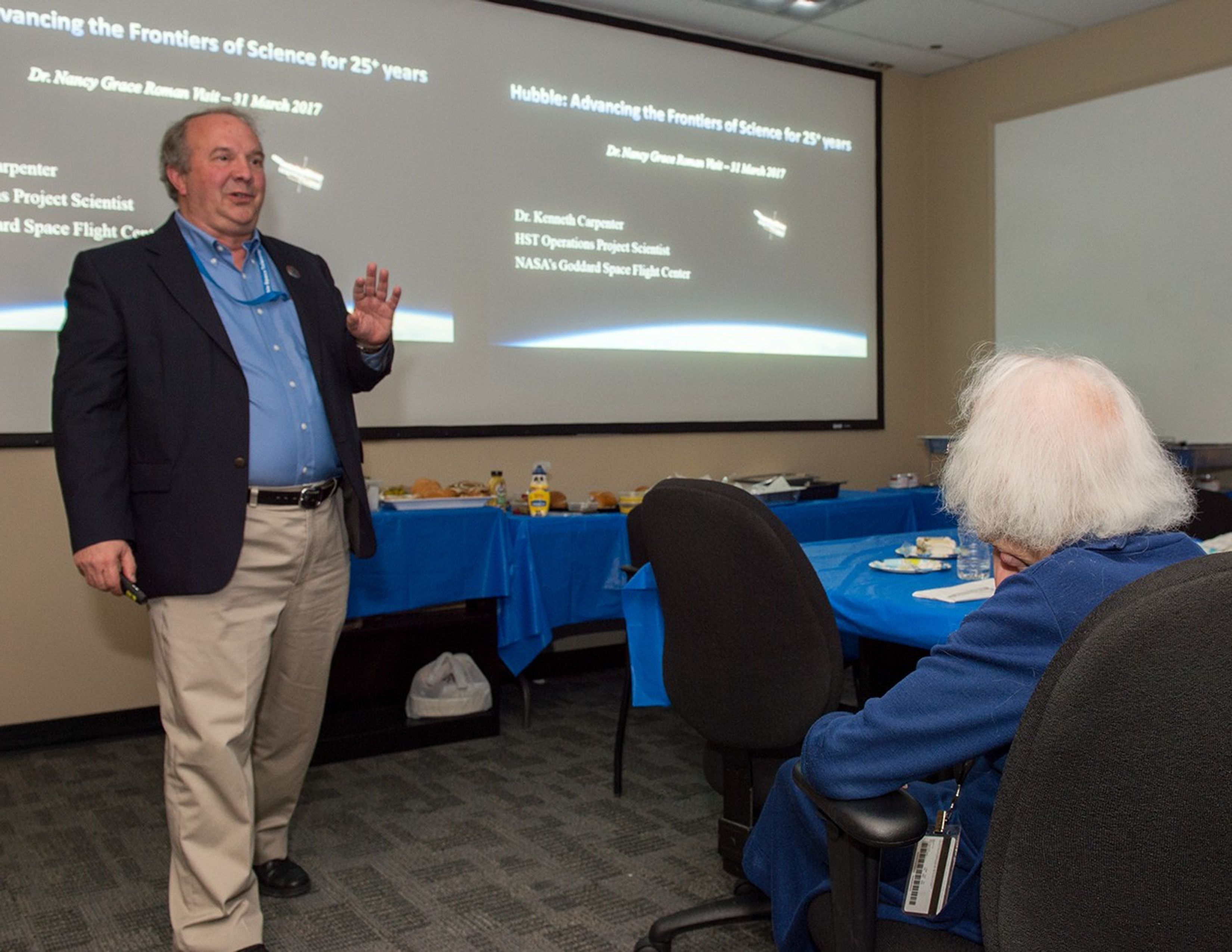 Dr. Roman, right, sits in a chair and faces away from the camera to look at Dr. Carpenter, left, and listen to him speak.