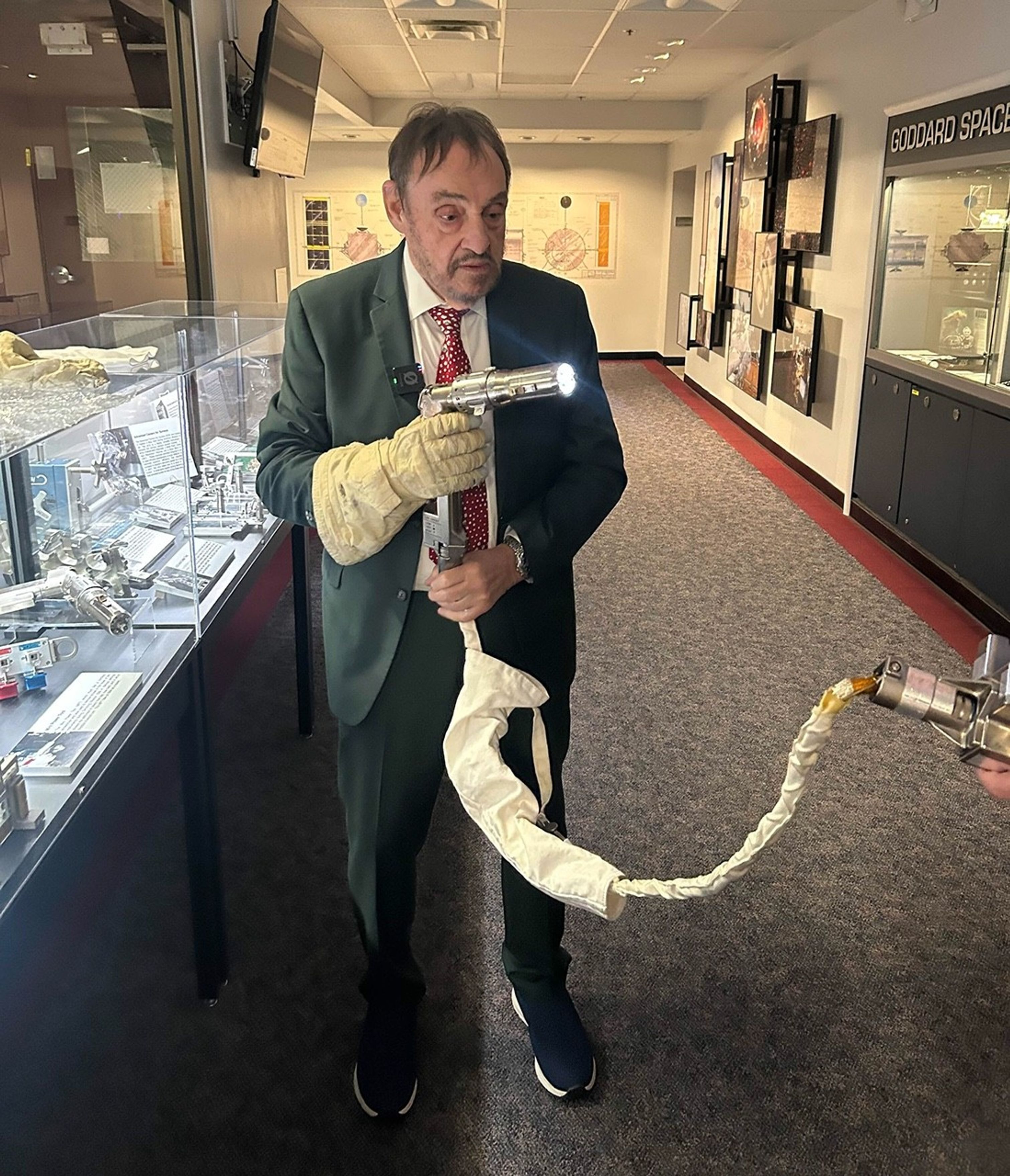 John Rhys-Davies wears an astronaut glove on his right hand and holds an astronaut power tool with a light on the end in both hands. He stands next to clear glass cases of astronaut tools on the left side of the image.