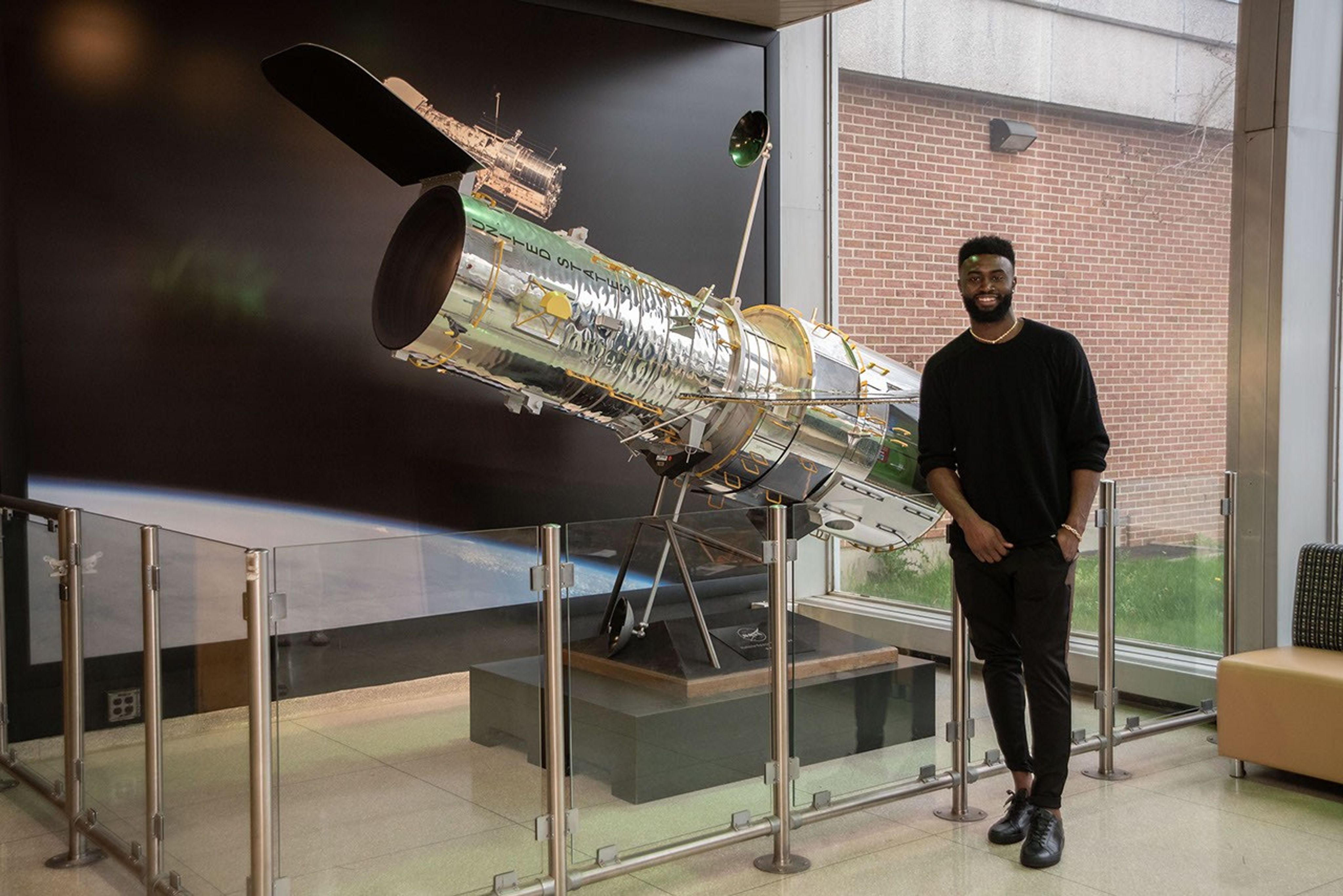 Jaylen Brown stands in front of a large scale model of Hubble, with him on the right and the model on the left.