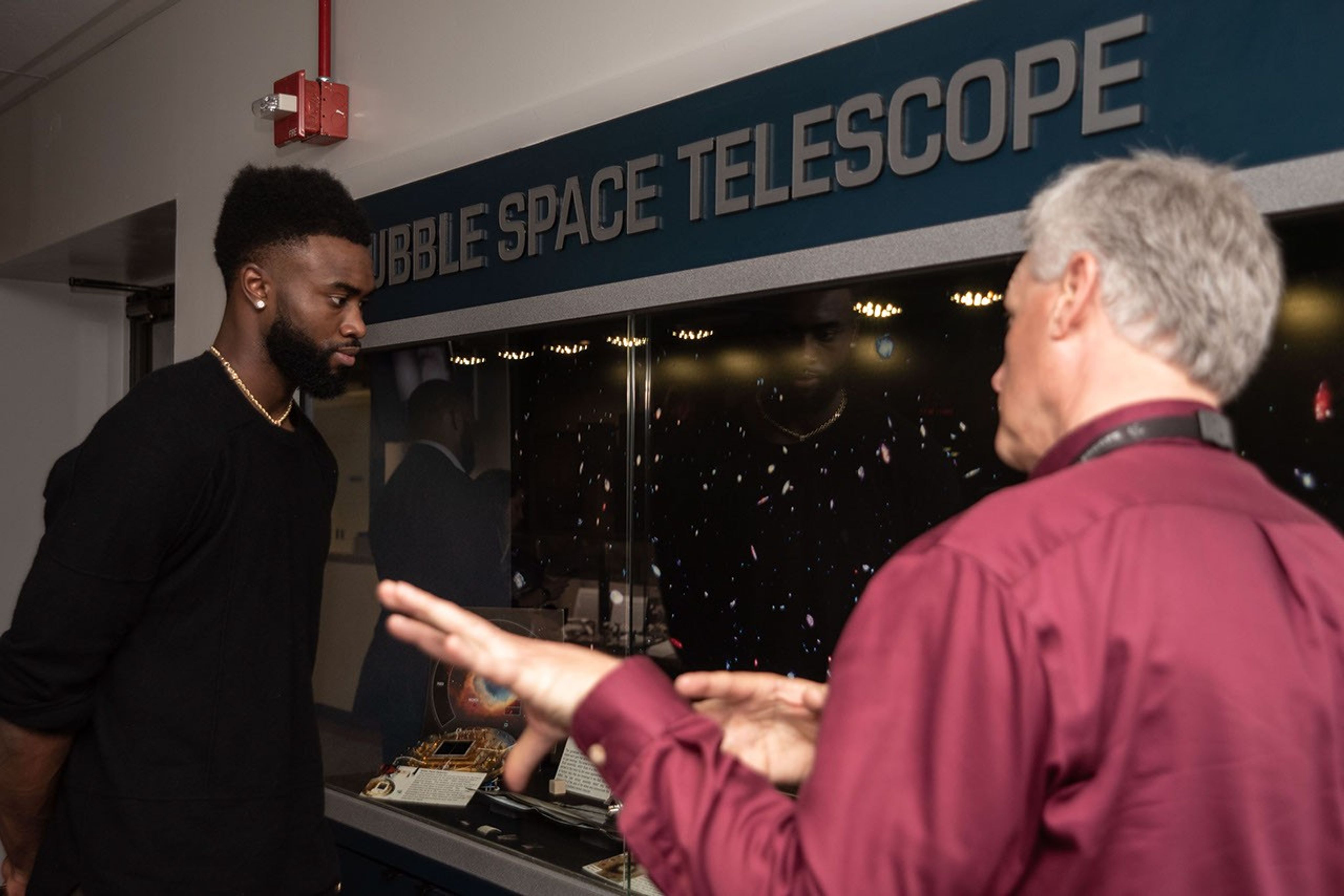 In the foreground, Jeletic (left) faces away from the camera toward Brown (left), speaking and motioning with his hands, while Brown examines Hubble instruments through the glass doors of the case.
