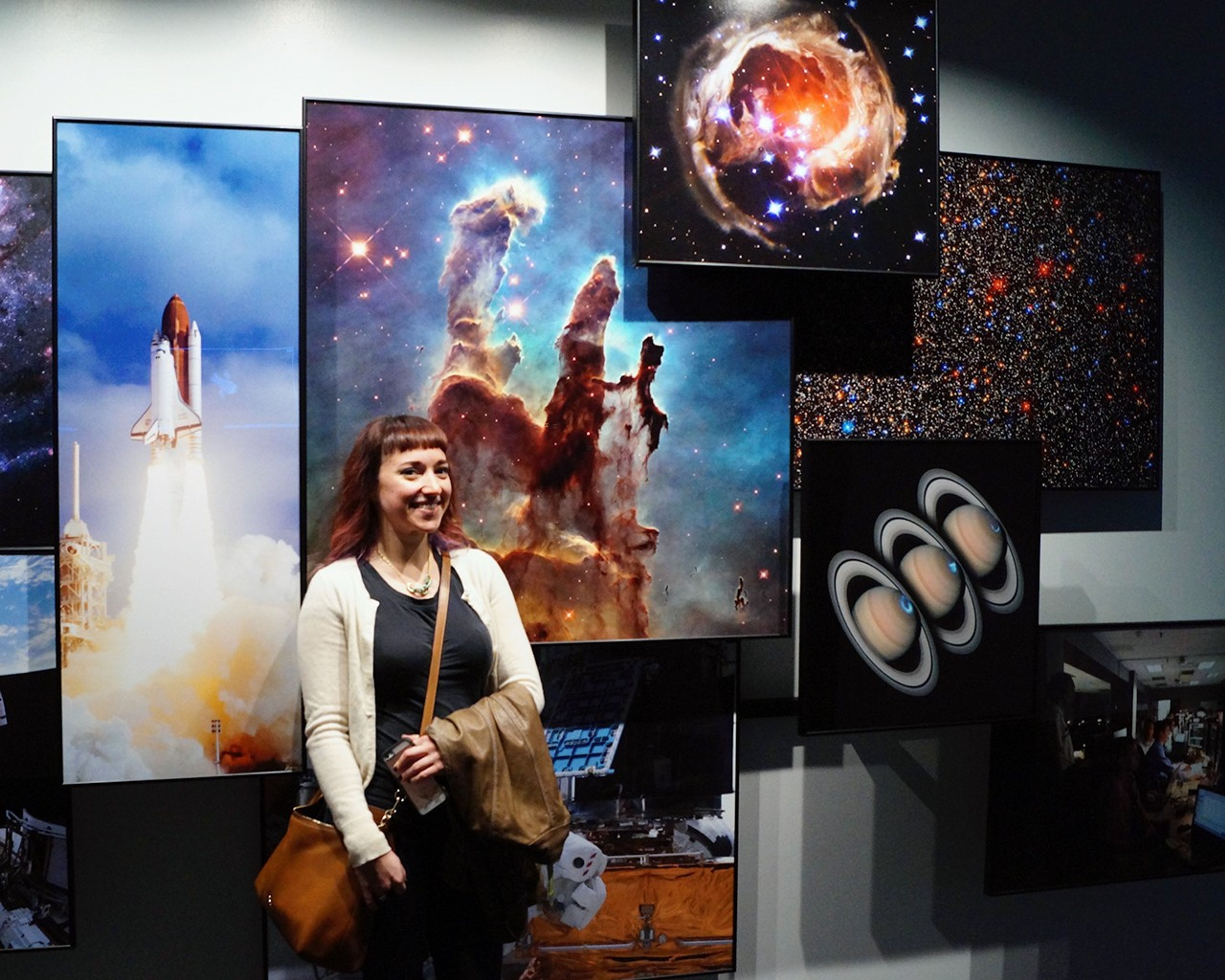 Amy Shira Teitel poses, smiling, in front of a montage of several Hubble images hung on a wall.