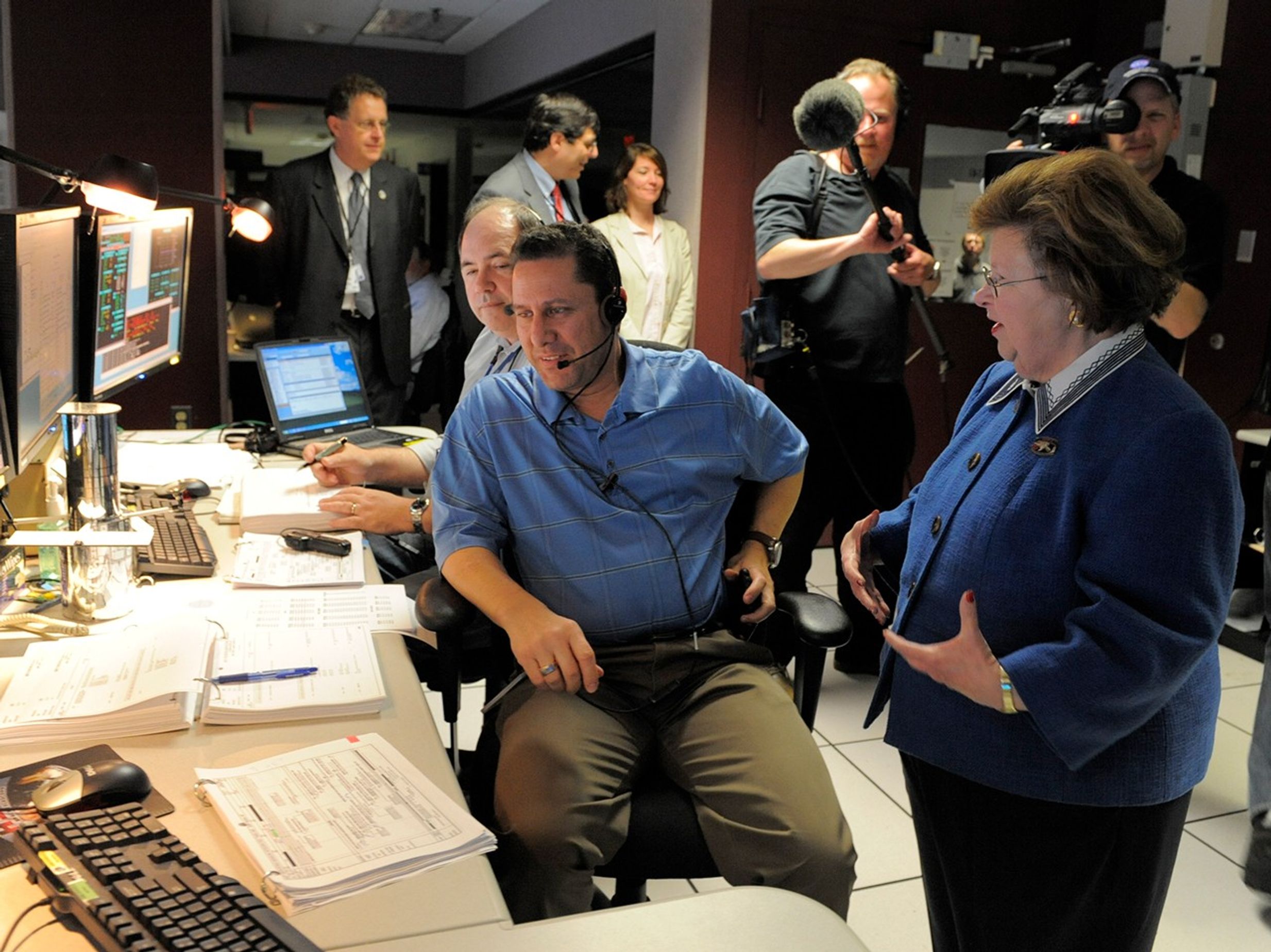 Senator Mikulski (right) speaks with two gentlemen sitting at a row of computers and monitors. Several more people are visible in the background, including two members of the camera crew (one with a camera and one with a microphone).