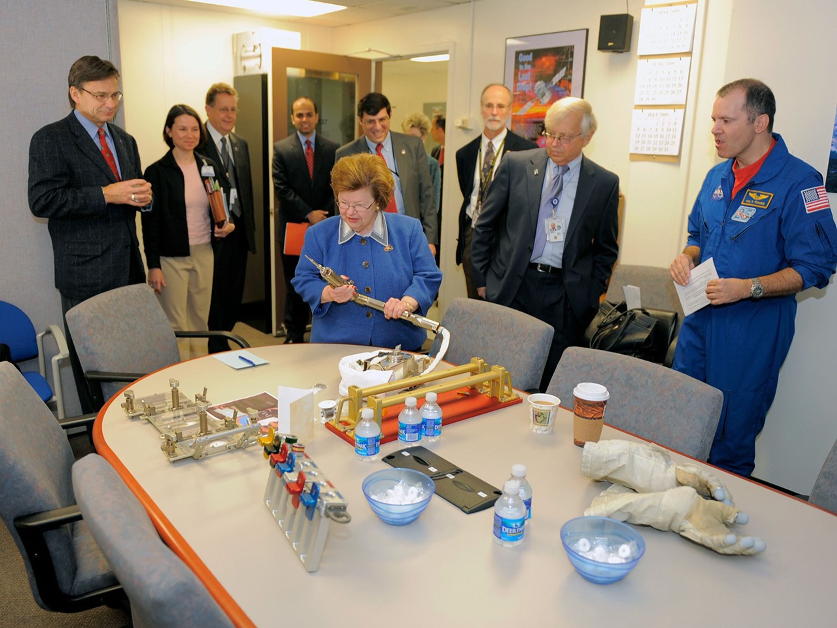 Senator Mikulski (center) stands at a conference roAdvom table holding a mini power tool in both hands, while eight smiling men and women watch, including an astronaut (farthest right). Also on the table are other astronaut tools, astronaut gloves, and bottles of water and bowls of mints.