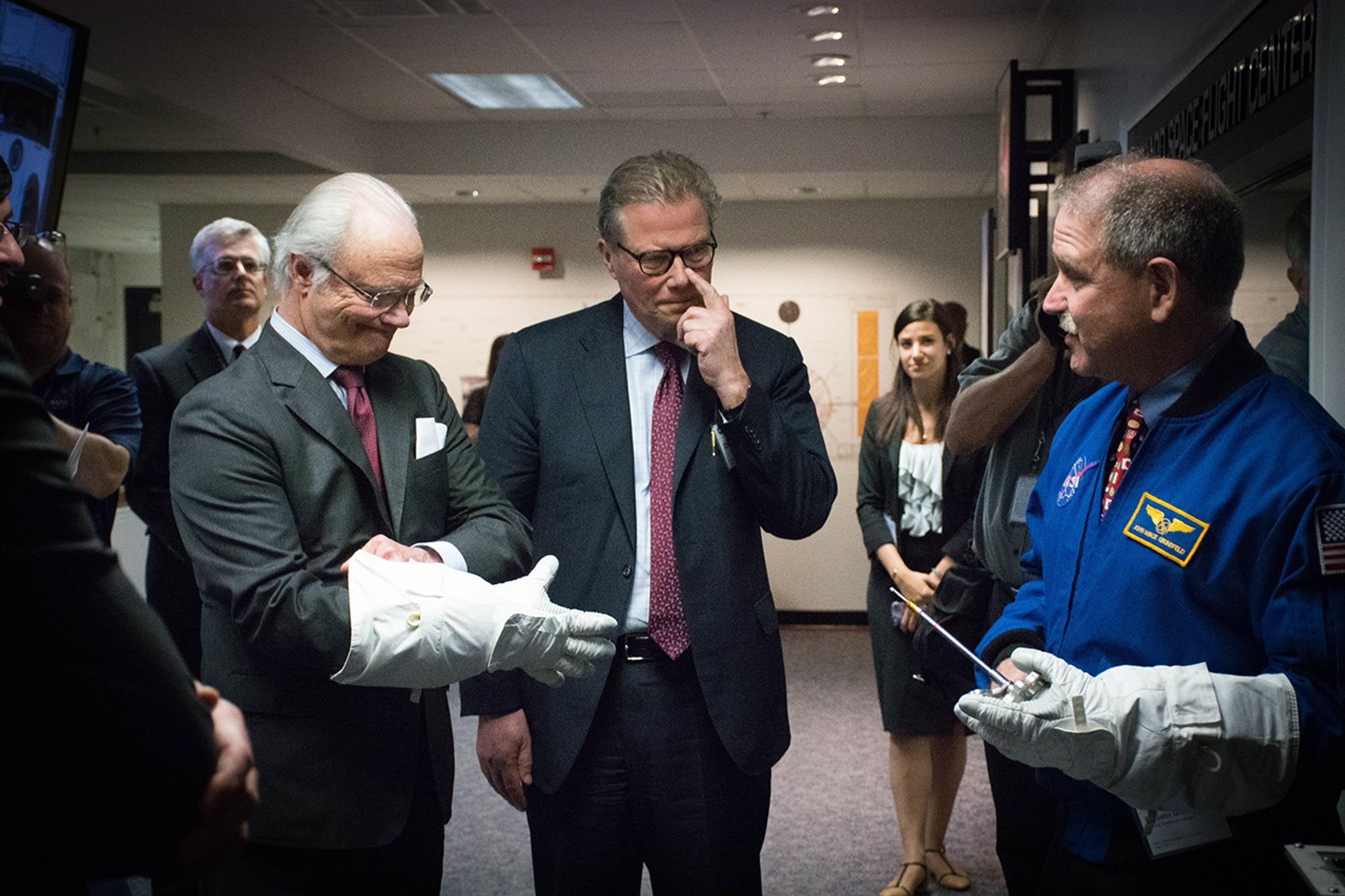 The King (left) pulls an astronaut glove onto his right hand, while another gentleman (center) stands beside him and Grunsfeld (right) wears a glove on his left hand while holding a tool and speaking to them. Several more people stand in the background watching.