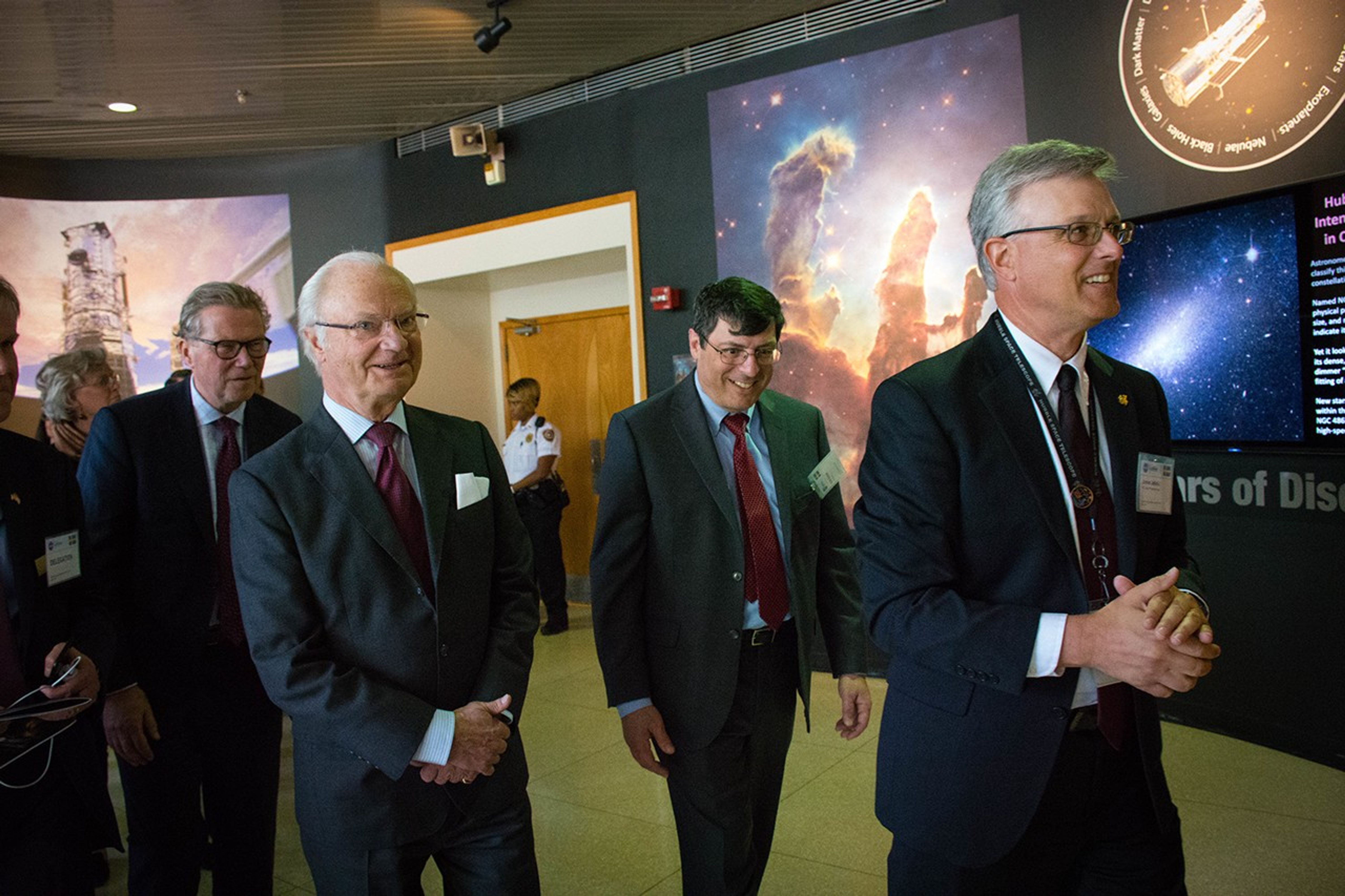 A group of men and women, led by Jeletic on the right, walk to the right side of the image, with the King in the center. In the background, a security police officer stands guard in front of a closed door.