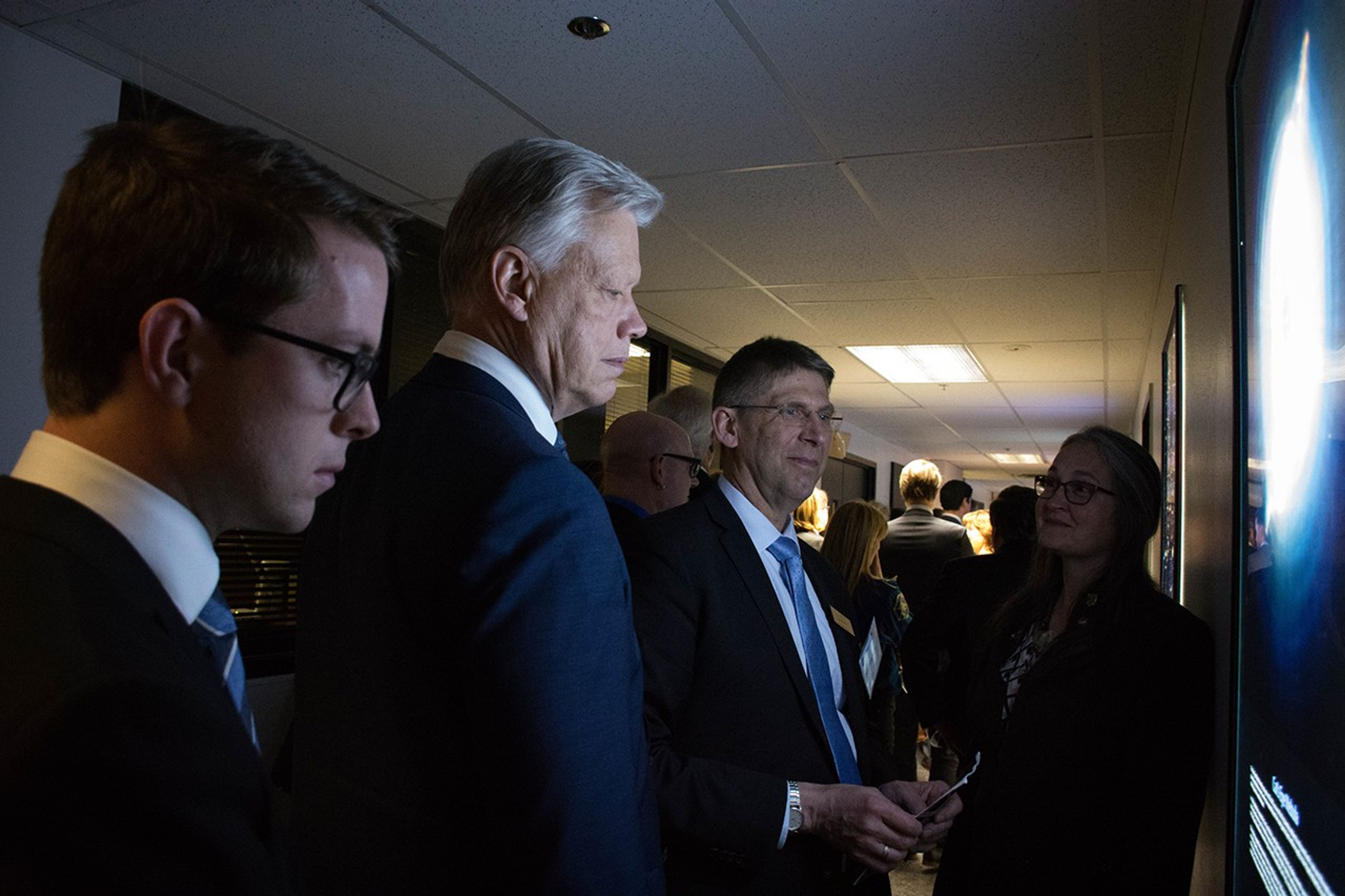 Padi Boyd, right, stands beside a lit-up image on the wall that she explains to three gentlemen looking at it. A large crowd of people is visible in the background, walking down the hall and facing away from the camera.