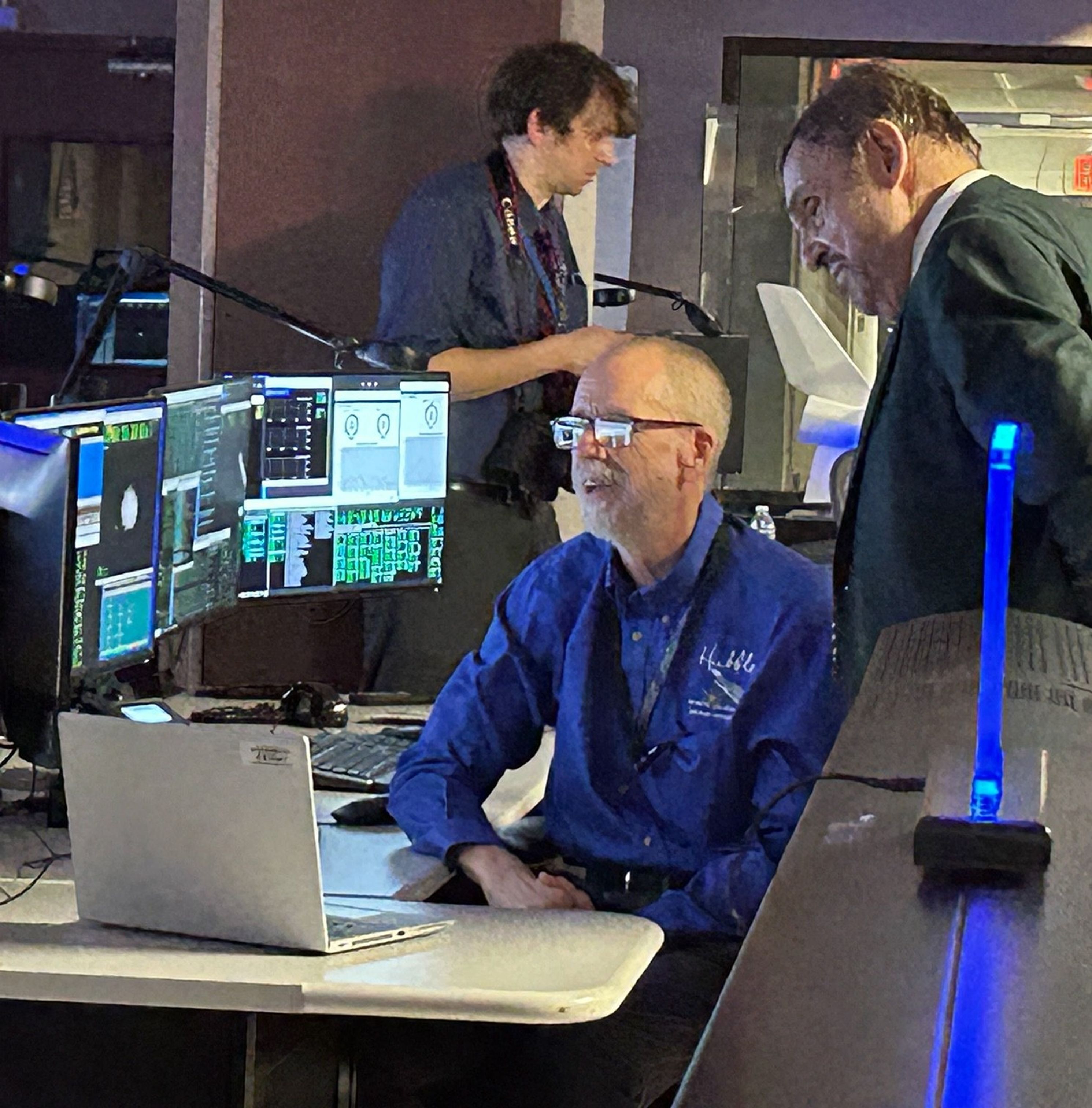 John Rhys-Davies (right) looks at the display of several monitors, at which Mike Wenz (center) is seated working. A photographer (left) is visible in the background.