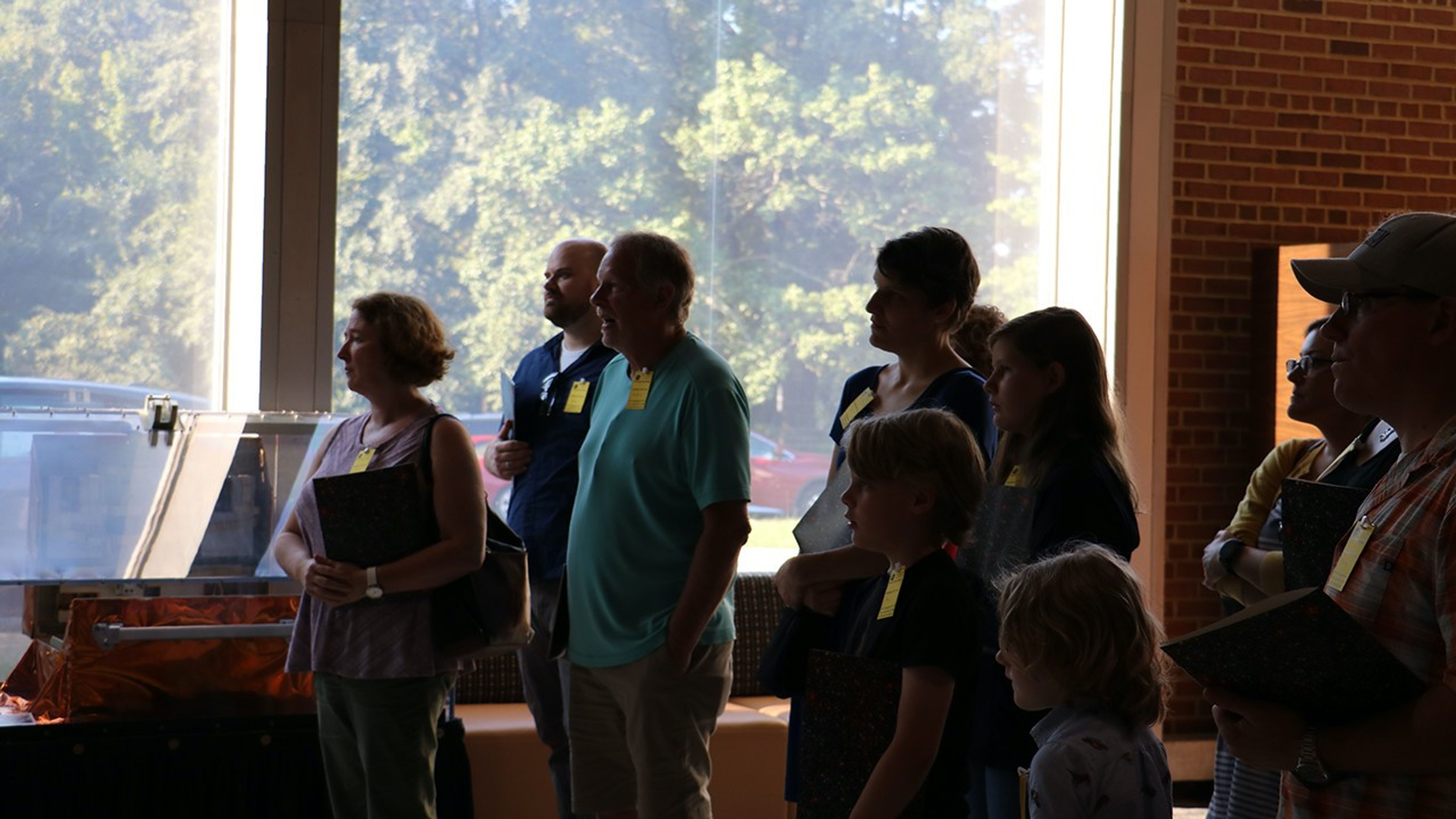 A crowd of adults and children (ten visible in this image) look to the left side of hte image while listening to a speaker (not shown) during a Hubble tour.
