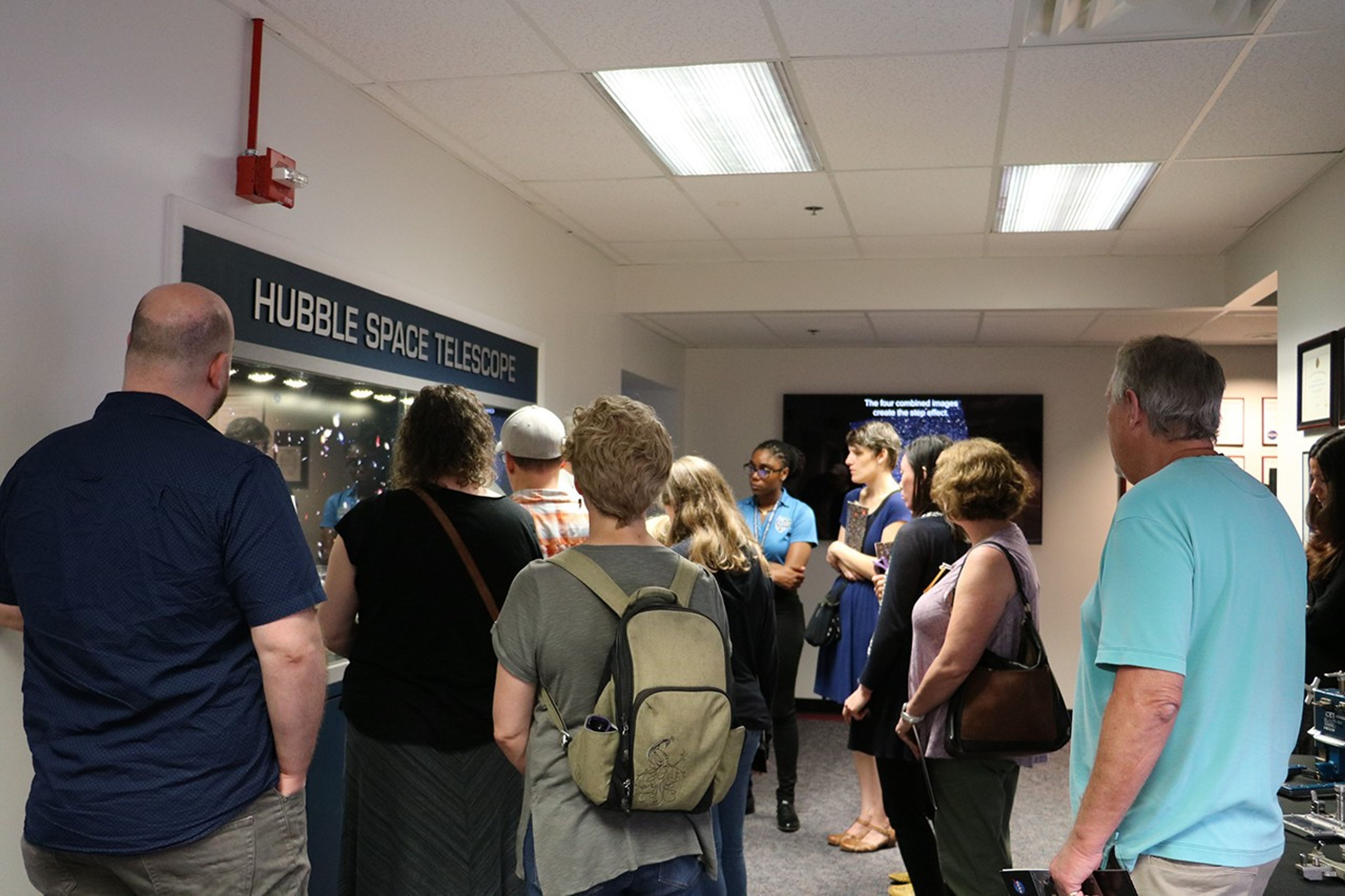 A group of adults and children (roughly 12 shown in this picture) face a Hubble display case of instruments, mostly facing away from the camera, during a tour.