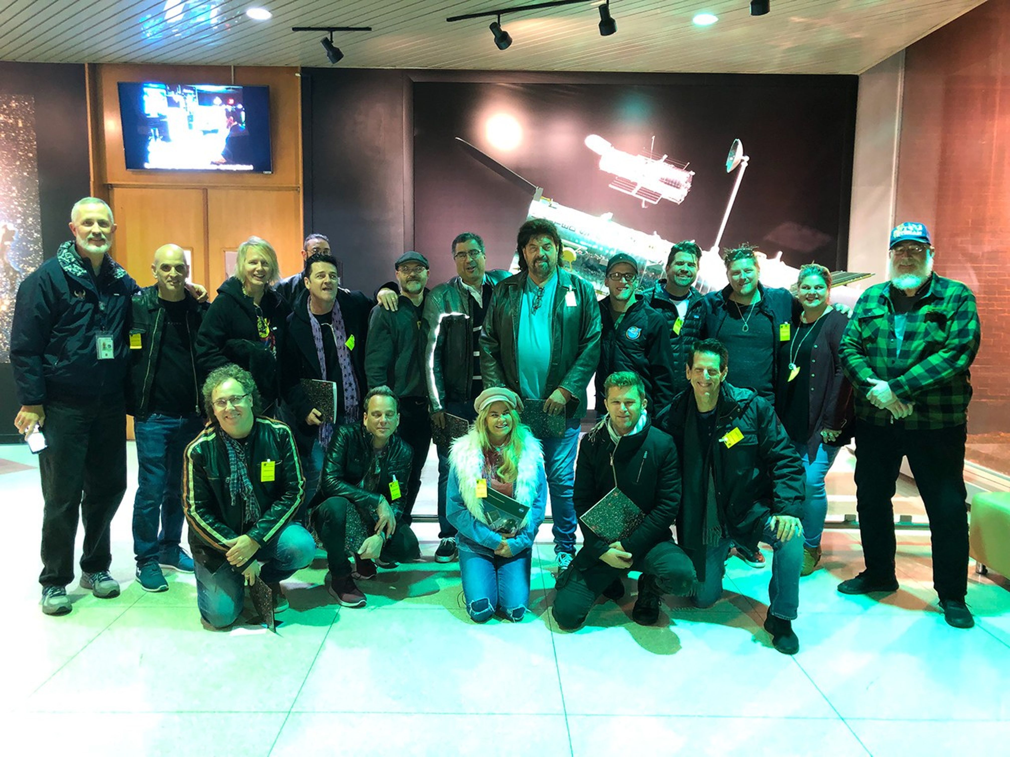 A group of 18 men and women smile and pose for a photo in front of a large model of Hubble. Five people kneel in a row in the front, while the remaining 13 stand in another row behind them.