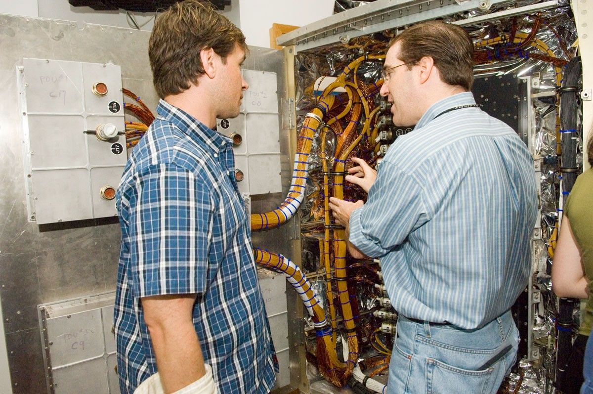 Cassidy, right, gestures toward the Power Control Unit trainer (a large box full of colorful wires, insulation material, and other electronic devices) while explaining it to a listening Trinneer, left.