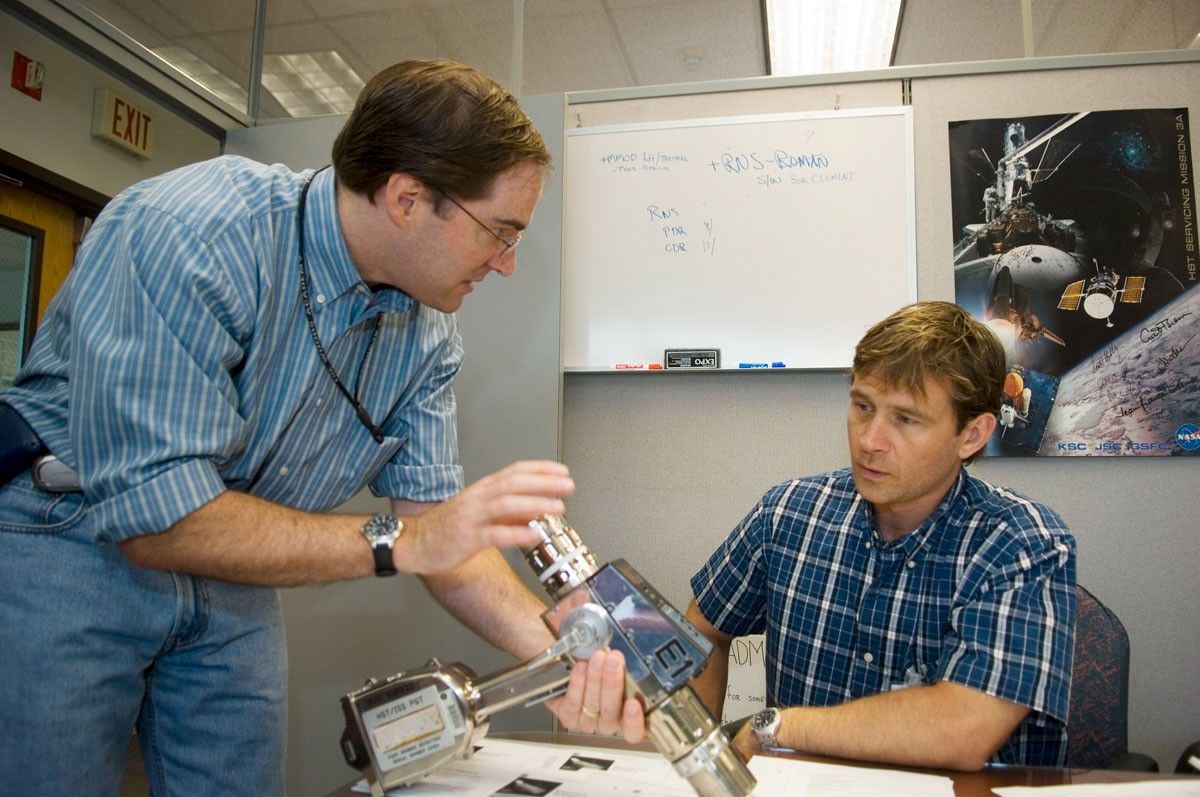 Cassidy, left, holds the pistol grip tool in his left hand while motioning with his right, and looks to Trinneer, right, seated at a table.