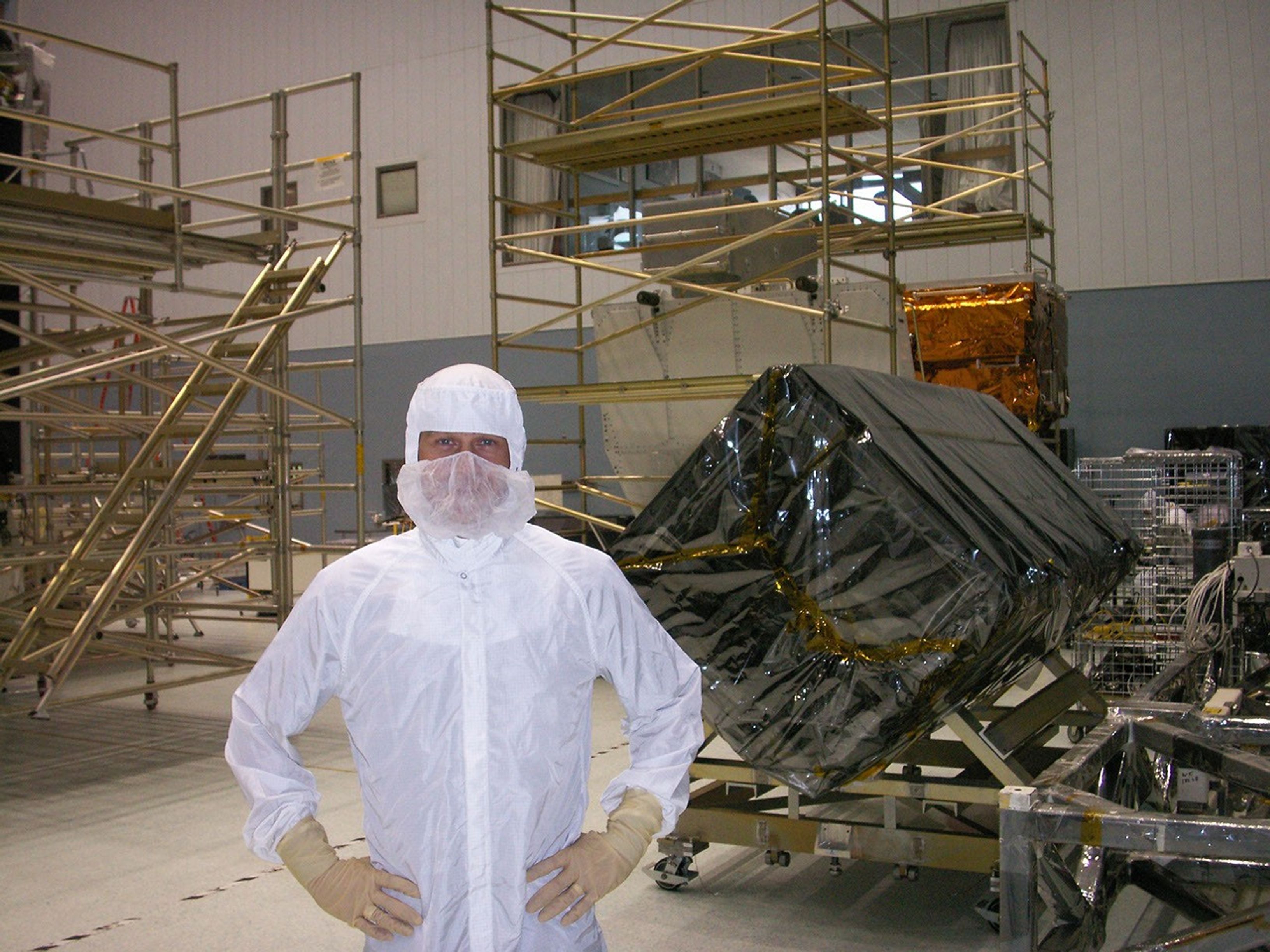 Trinneer, wearing a white clean suit with a mask over the face, stands with his hands on his hips in front of an instrument wrapped in translucent black material with a square base and rectangular sides. In the background in the center and left are several gold railings and ladders.