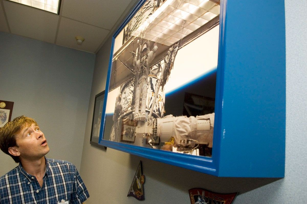 Trinneer, left, gazes up into a photo of an astronaut servicing mission, mounted on the wall in a blue box.