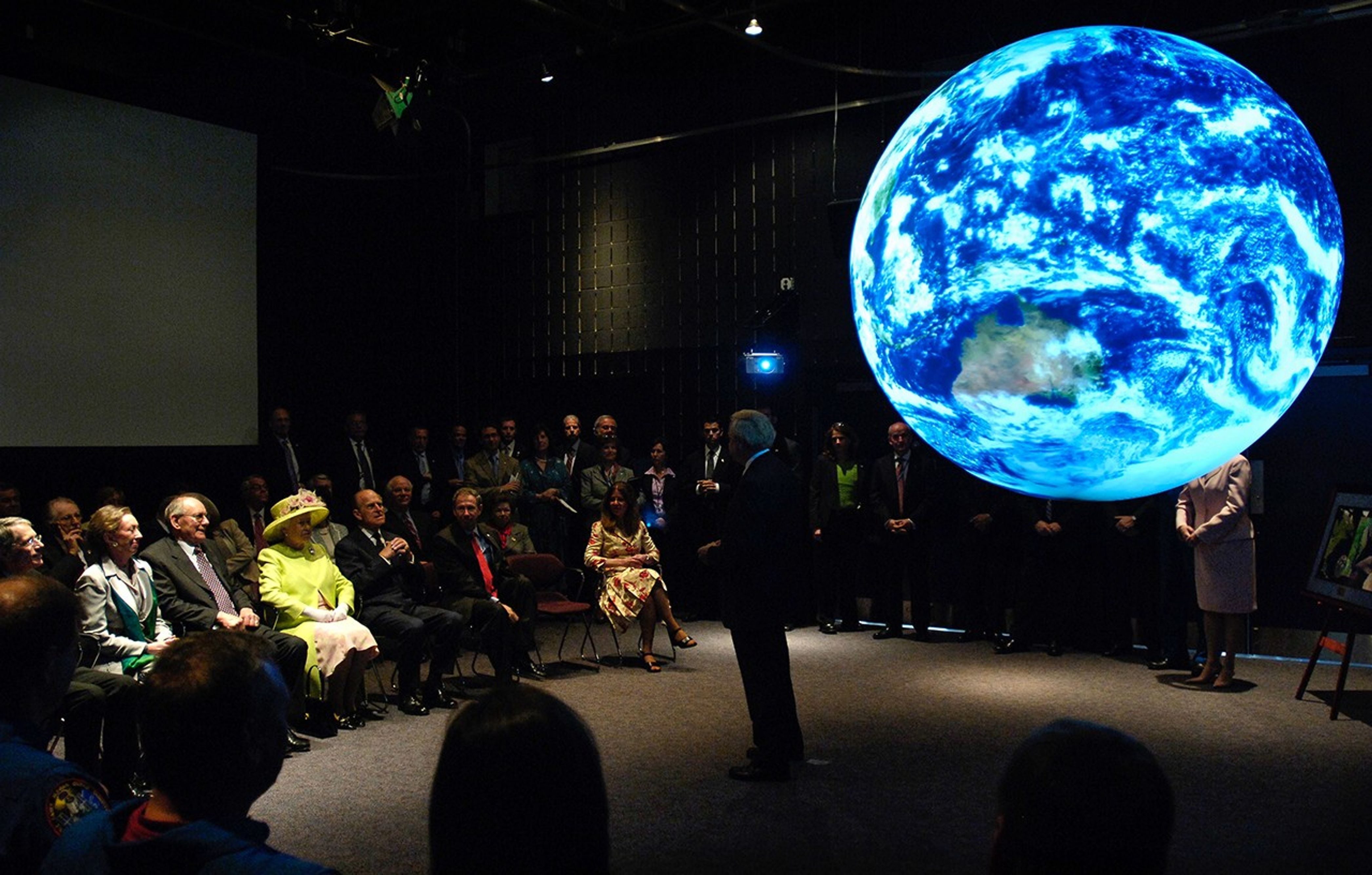 On the right, a image of the Earth is projected onto a sphere suspended above the floor. In the center, a male presenter stands in front of the sphere while speaking to the audience on the left side of the image, including the Queen and Prince.
