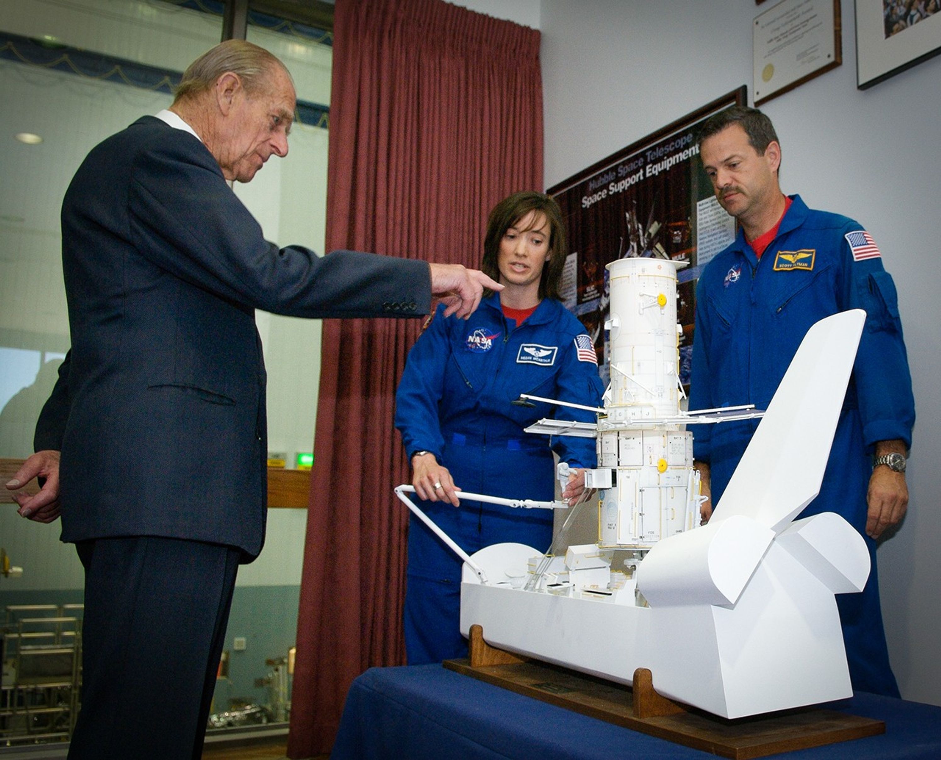 Astronauts Altman (right) and McArthur (center) explain the model of Hubble and the space shuttle to Prince Philip (left), who points at it.