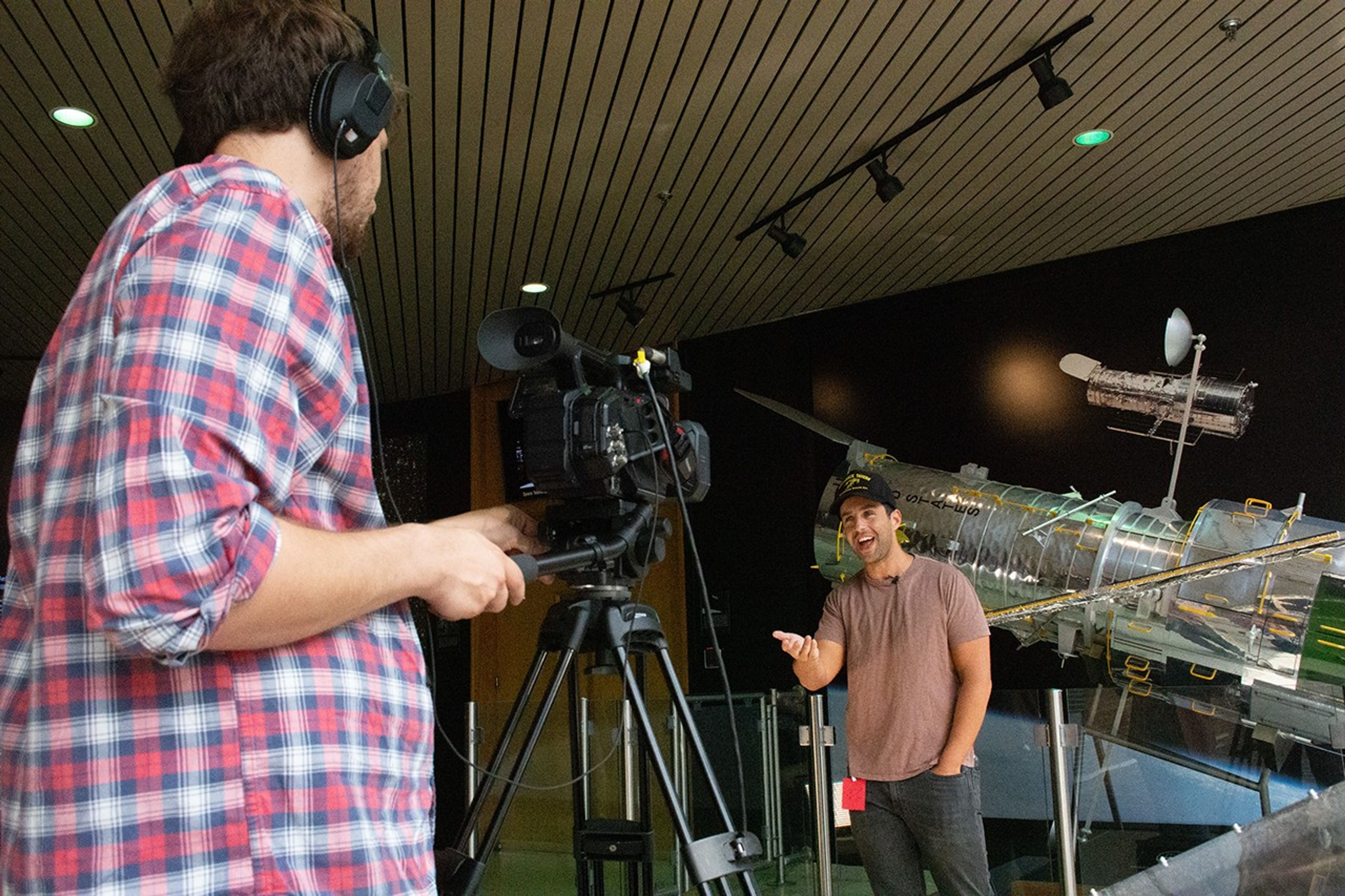 Josh Peck stands in front of a large scale model of Hubble while speaking to and motioning with his hand toward a video camera that, in the foreground, Paul Morris is operating while facing away from the camera that took this picture.