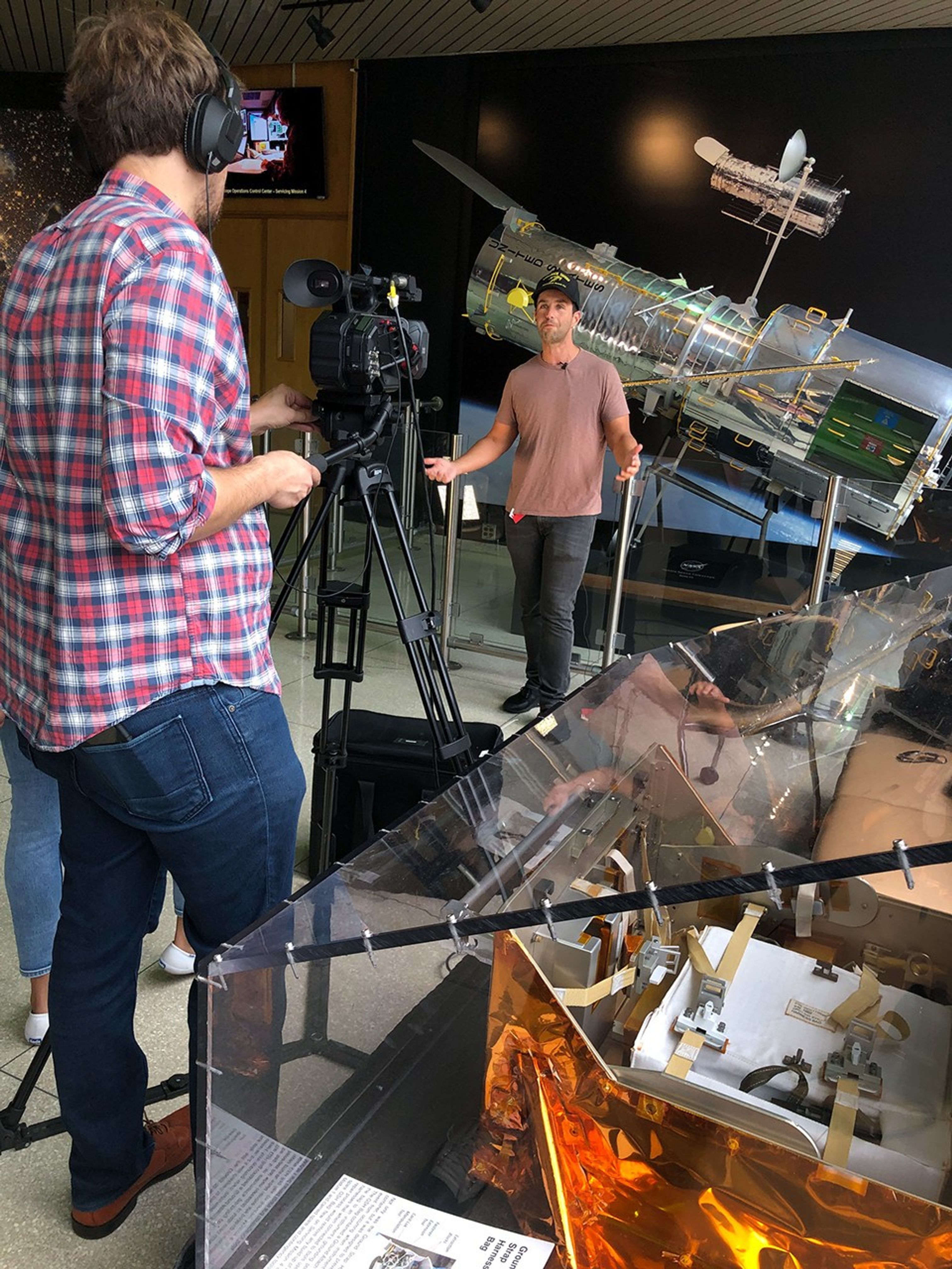 Josh Peck stands in front of a large scale model of Hubble with his hands outspread while speaking to a video camera that, in the foreground, Paul Morris is operating while facing away from the camera that took this picture. To his right, also in the foreground, is a clear glass case containing astronaut tools.