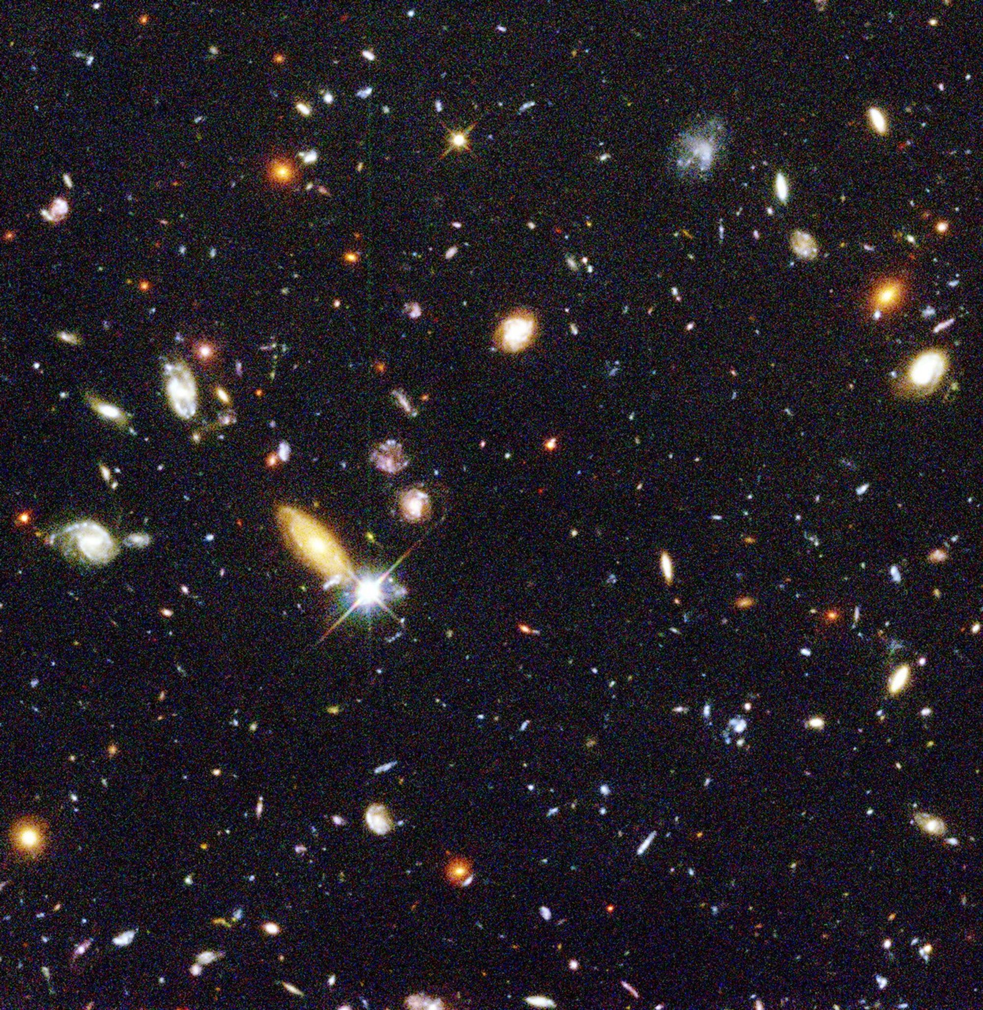 On a grainy black background, dozens of galaxies of various shapes, sizes, and colors are scattered.