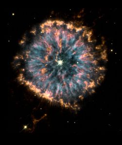 A cloud of gas and dust resembling an eye. The outer edges of the eye are pale orange; the "iris" is blue, orange, and pink; and the center "pupil" is darker and contains a glowing star. The background is black, with faint stars in the background and wisps of orange gas.
