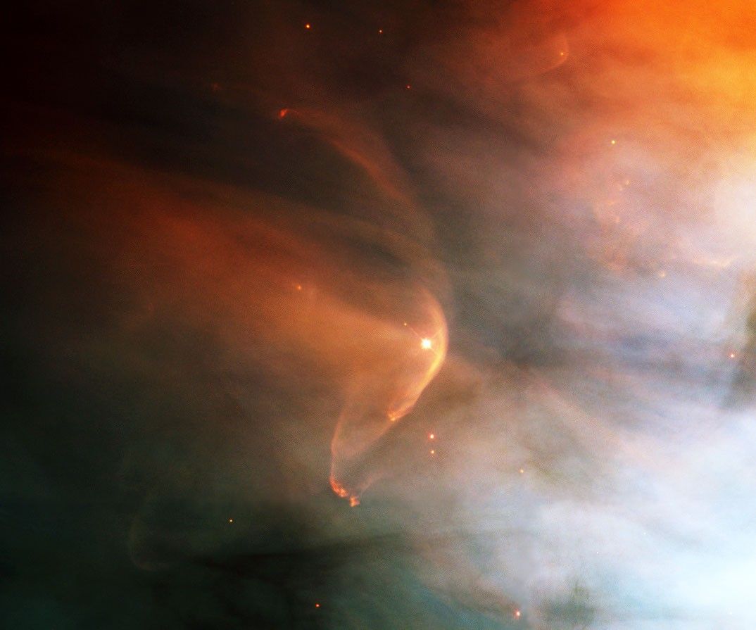 A very small portion of a larger nebula with a distinct arc of yellow-orange and more diffuse orange gas streaming away to its upper left.