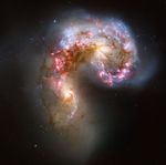 Visible light emitted by the Antennae galaxies is shown in various colors, including pink, blue, brown, and white. The overall shape of the two colliding galaxies resembles a curled-up shrimp, with one of the galaxy’s main spiral arm toward the top right and the other galaxy’s main spiral arm toward the bottom left.