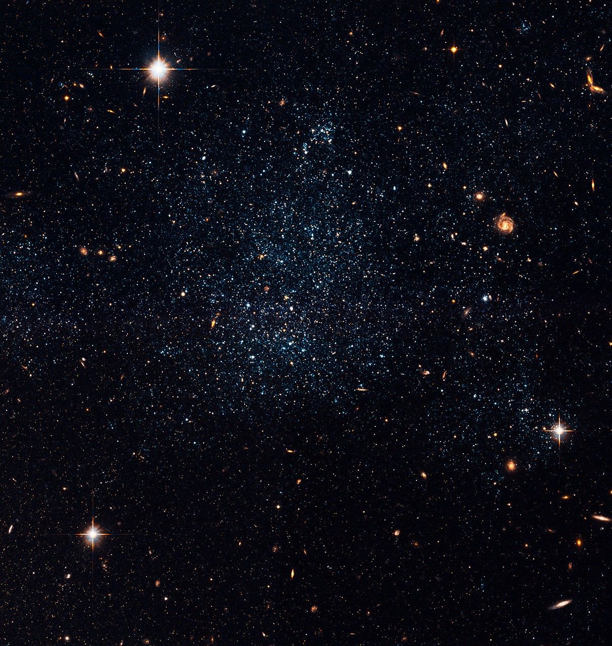 A field of blue, white, and red stars and galaxies against the black background of space. There is an irregular, denser grouping of stars in the center.