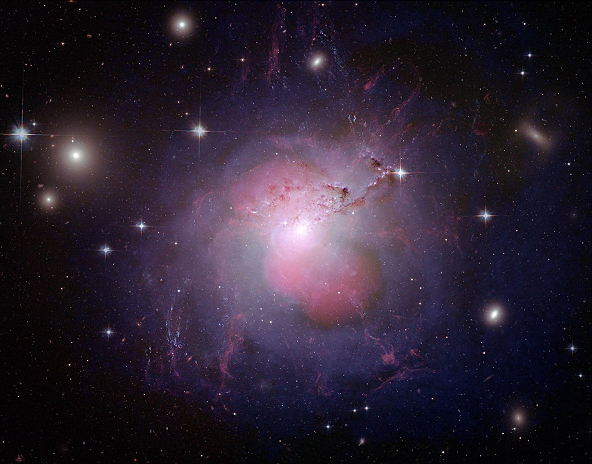 A galaxy with a white center surrounded by pink lobes above and below it. Outside the pink lobes is a violet shell that surrounds the galaxy. From the center of the galaxy are stringy filaments of red gas.