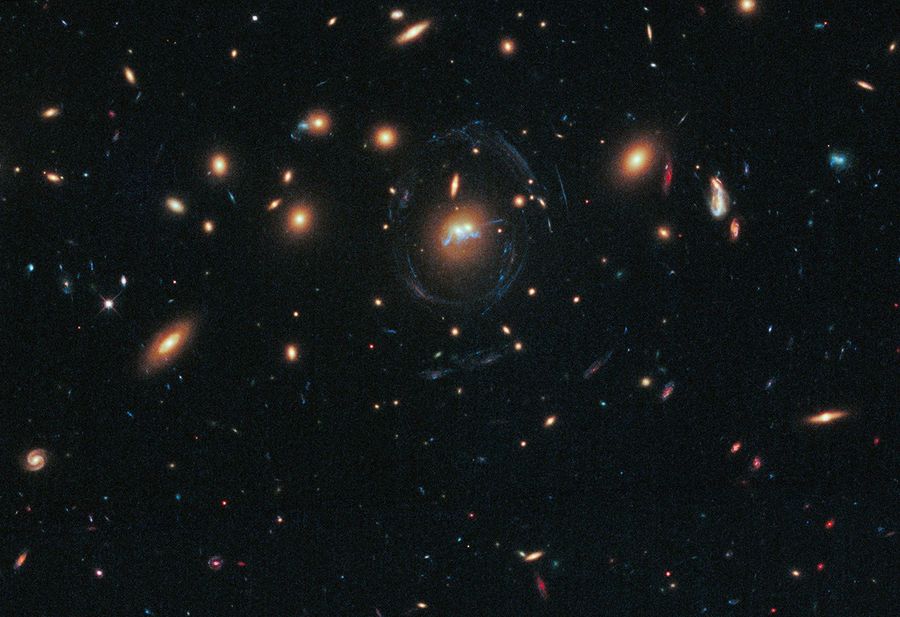 Black background dotted with galaxies. Two reddish galaxies at image center as two dots next to each other, with a thin bluish-white ring curving almost completely around it. Directly below the two galaxies, the filaments appear as a squiggly blue line.