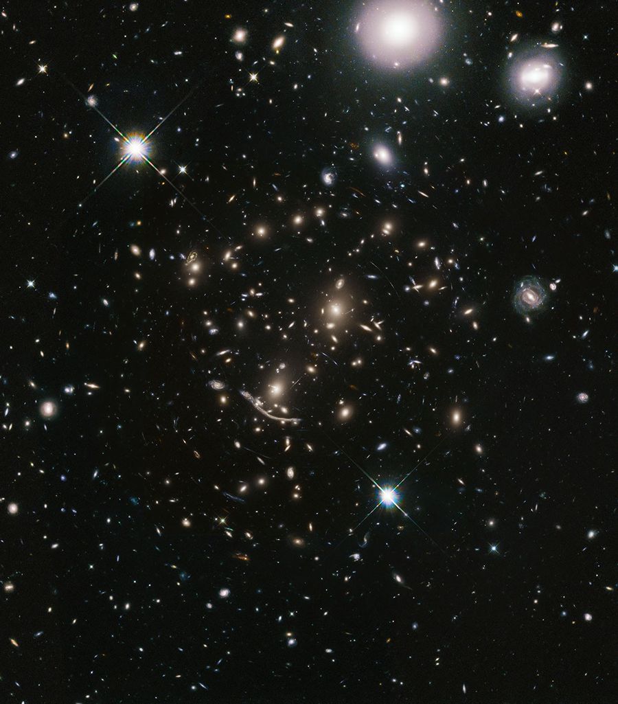 A packed field of galaxies and curved blue streaks and arcs. Large grouping of galaxies warped around what appears to be a giant lens. Each galaxy appears as a glob of light. Bright blue-white foreground star at lower-center and upper left, and bright white foreground galaxies at upper-right.