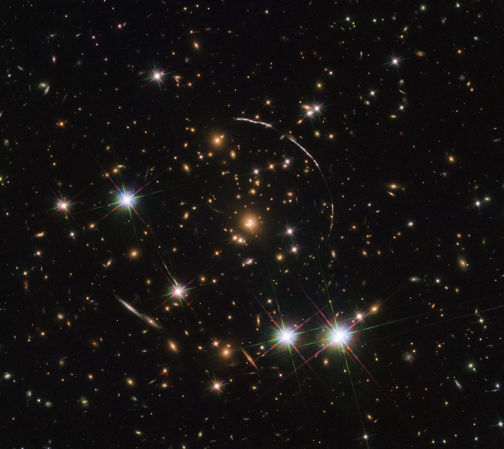 A field full of distant galaxies on a dark background. Most of the galaxies appear small, but there are a few larger galaxies and some stars where detail is visible. In the very center is a brightly-glowing red elliptical galaxy and a broad disk. A whitish, warped, ring of light--a large arc in the upper-right and a smaller one in the lower-left- surrounds its core at a distance. Several bright stars are visible with the "crosshair" patterns across them, two beside each other on the bottom-right, another on the left, and a few more smaller ones scattered around.