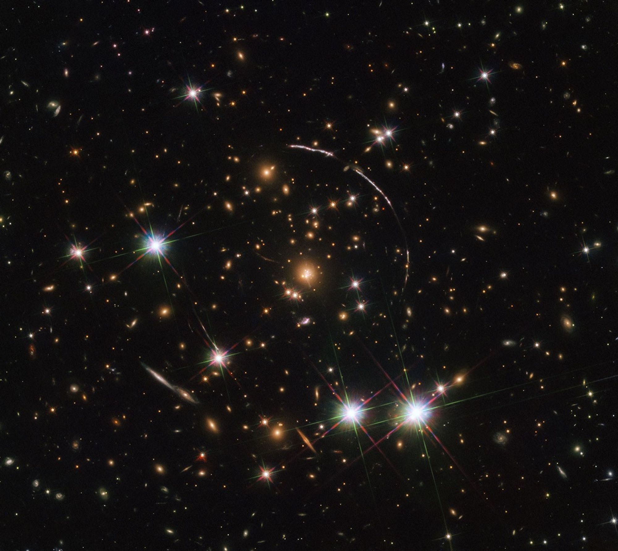 A field full of distant galaxies on a dark background. Most of the galaxies appear small, but there are a few larger galaxies and some stars where detail is visible. In the very center is a brightly-glowing red elliptical galaxy and a broad disk. A whitish, warped, ring of light--a large arc in the upper-right and a smaller one in the lower-left- surrounds its core at a distance. Several bright stars are visible with the "crosshair" patterns across them, two beside each other on the bottom-right, another on the left, and a few more smaller ones scattered around.