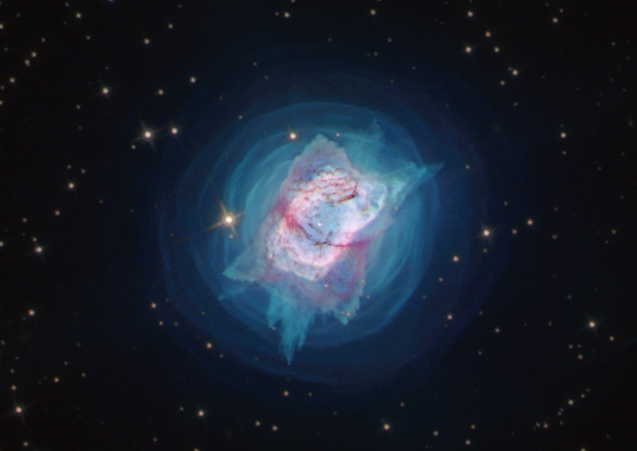 A nebula with a pink elliptical shell is surrounded by concentric rings of blue gas, all set on a black star-filled background.