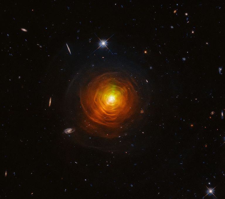 Red-orange arcs surround the carbon star CW Leonis. Brilliant searchlight beams from the star’s surface poke through.