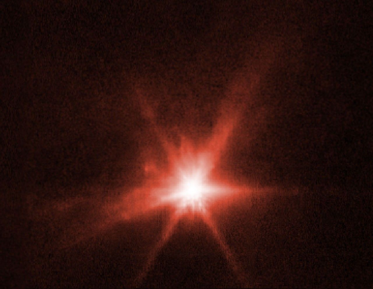 On a black background, a bright red spot appears at the center of the image. The spot, which is the asteroid Didymos-Dimorphos system after impact from DART, has 8 diffraction spikes extending out from its center. Also surrounding the asteroid is a haze of bright light with wispy tendrils extending outwards.