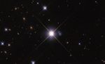 A large bright star is centered, with four long rays extending in an X shape. Between the rays on the right is a small blue peanut-shaped galaxy with some bright spots. Smaller similar stars and orange background galaxies fill image.