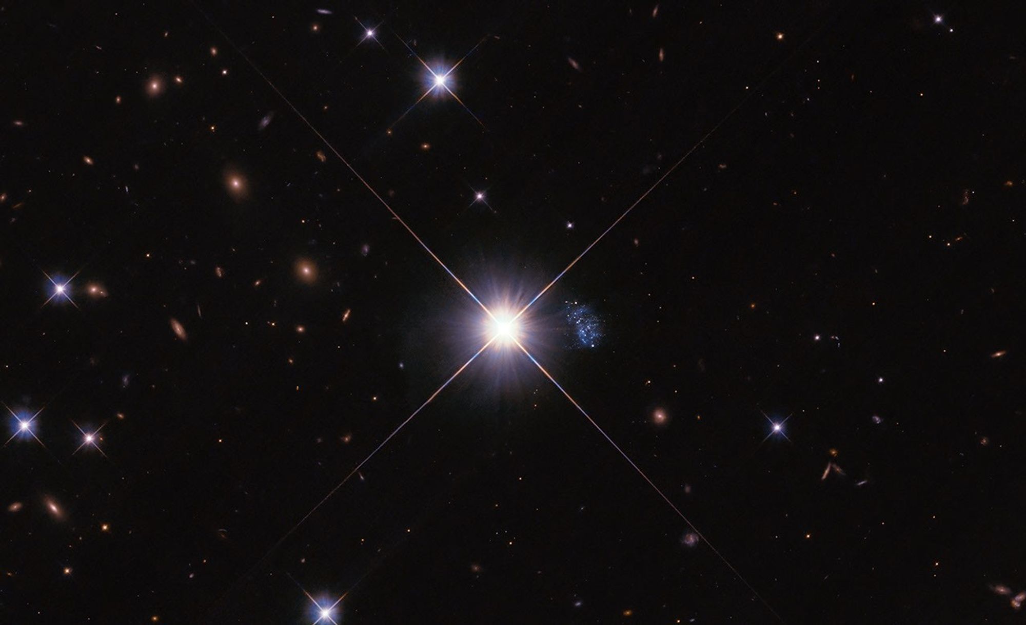 A large bright star is centered, with four long rays extending in an X shape. Between the rays on the right is a small blue peanut-shaped galaxy with some bright spots. Smaller similar stars and orange background galaxies fill image.