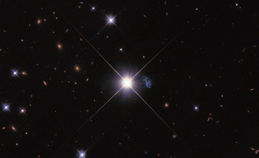 A large bright star is centered, with four long rays extending in an X shape. Between the rays on the right is a small blue peanut-shaped galaxy with some bright spots. Smaller similar stars and orange background galaxies fill image.