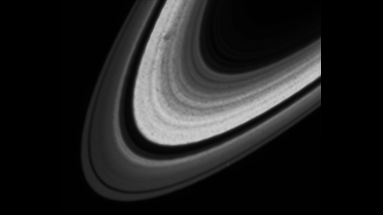 Series of black and white close-up images on the curve of Saturn's rings, with no sphere of the planet visible. Images together create a GIF video showing dark smudges appearing to move along the bright rings of Saturn. One dark area remains distinct throughout, while the other goes in and out of distinction and darkness.