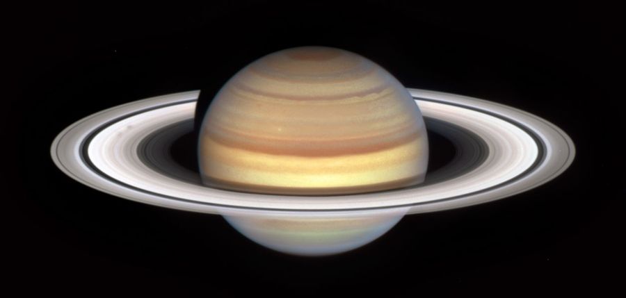 Planet Saturn with bright white rings and multi-colored main sphere. Spoke features appear like faint gray smudges against the rings' bright backdrop, about midway from the planet to the rings' outer edge. Above the rings plane, the planet's bands are shades of red and orange, with brighter yellow nearer the equator.
